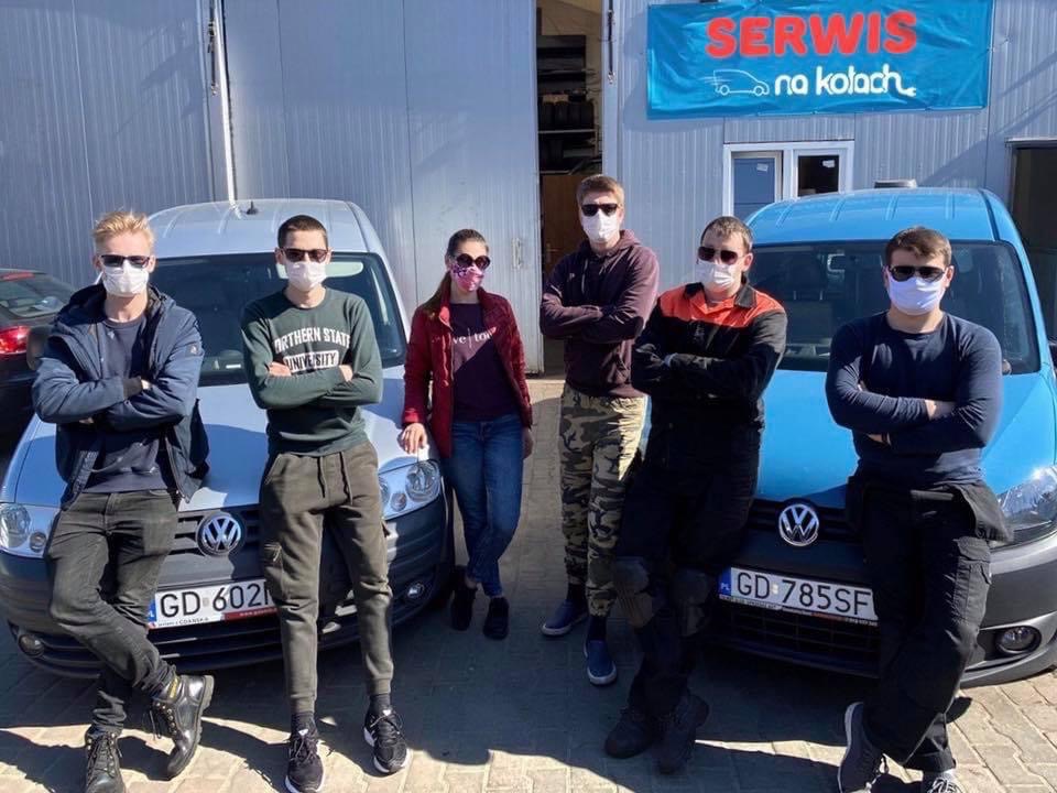 Zespół serwisowy w maskach ochronnych stoi przed dwoma samochodami dostawczymi Volkswagen, srebrnym i niebieskim, z tabliczką 'Serwis na kołach' widoczną w tle.