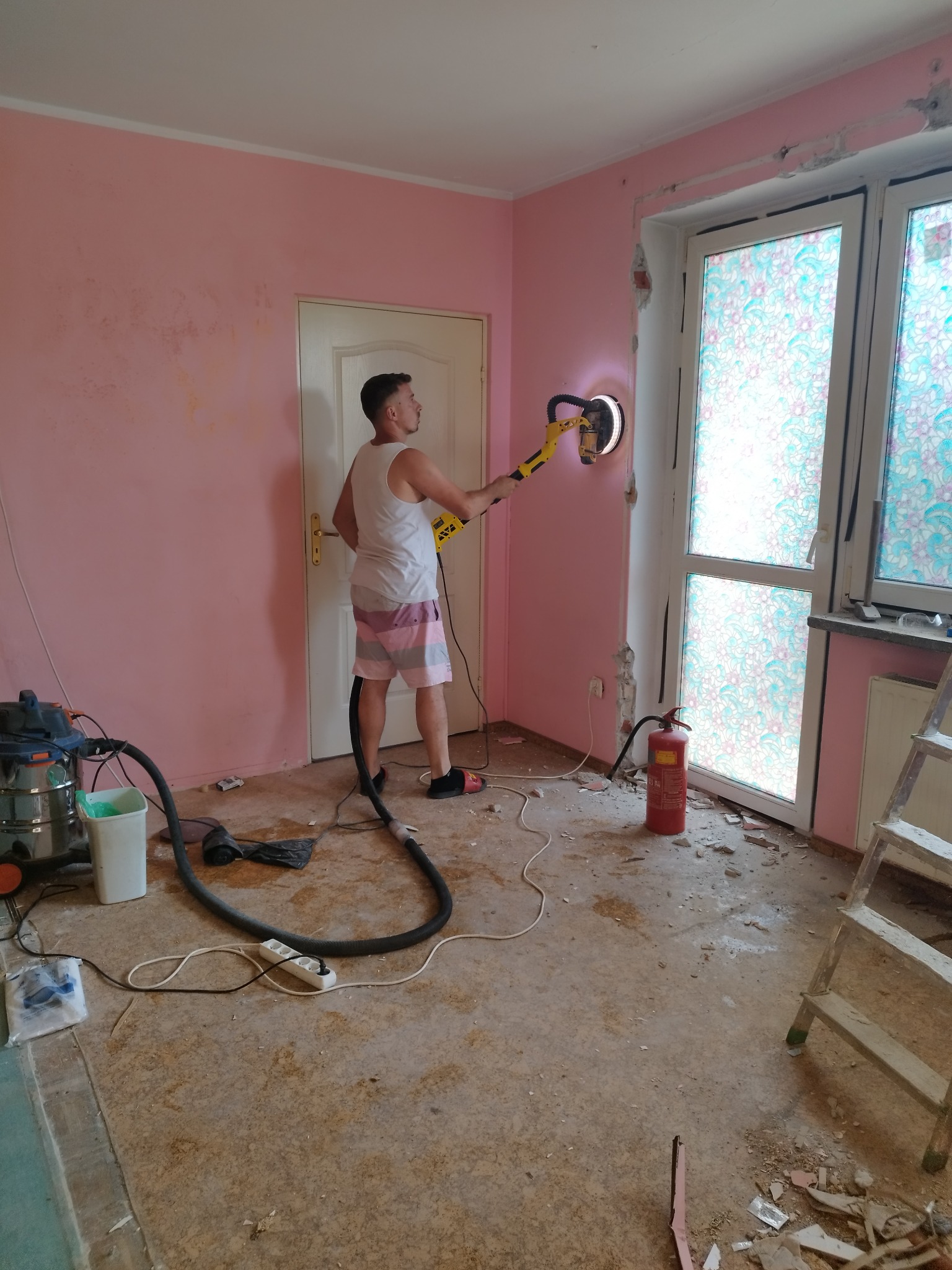Mężczyzna w krótkich spodenkach szlifuje ścianę szlifierką oscylacyjną w pokoju z różowymi ścianami podczas remontu. Widoczny odkurzacz przemysłowy, drabina i gaśnica.