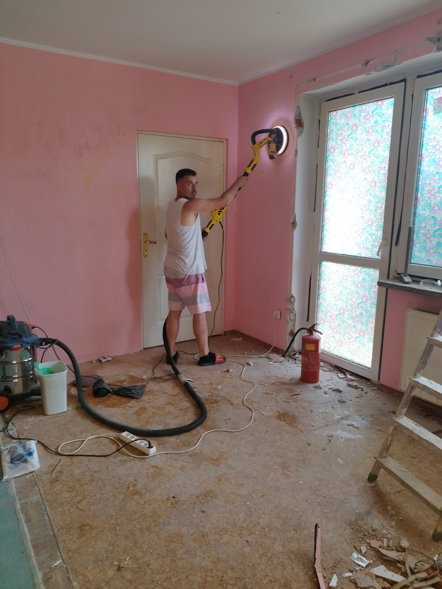 Mężczyzna w krótkich spodenkach szlifuje ścianę w pokoju o różowych ścianach za pomocą szlifierki z odkurzaczem; widoczne ślady po usuniętych listwach przypodłogowych, drabina, gaśnica i śmieci...