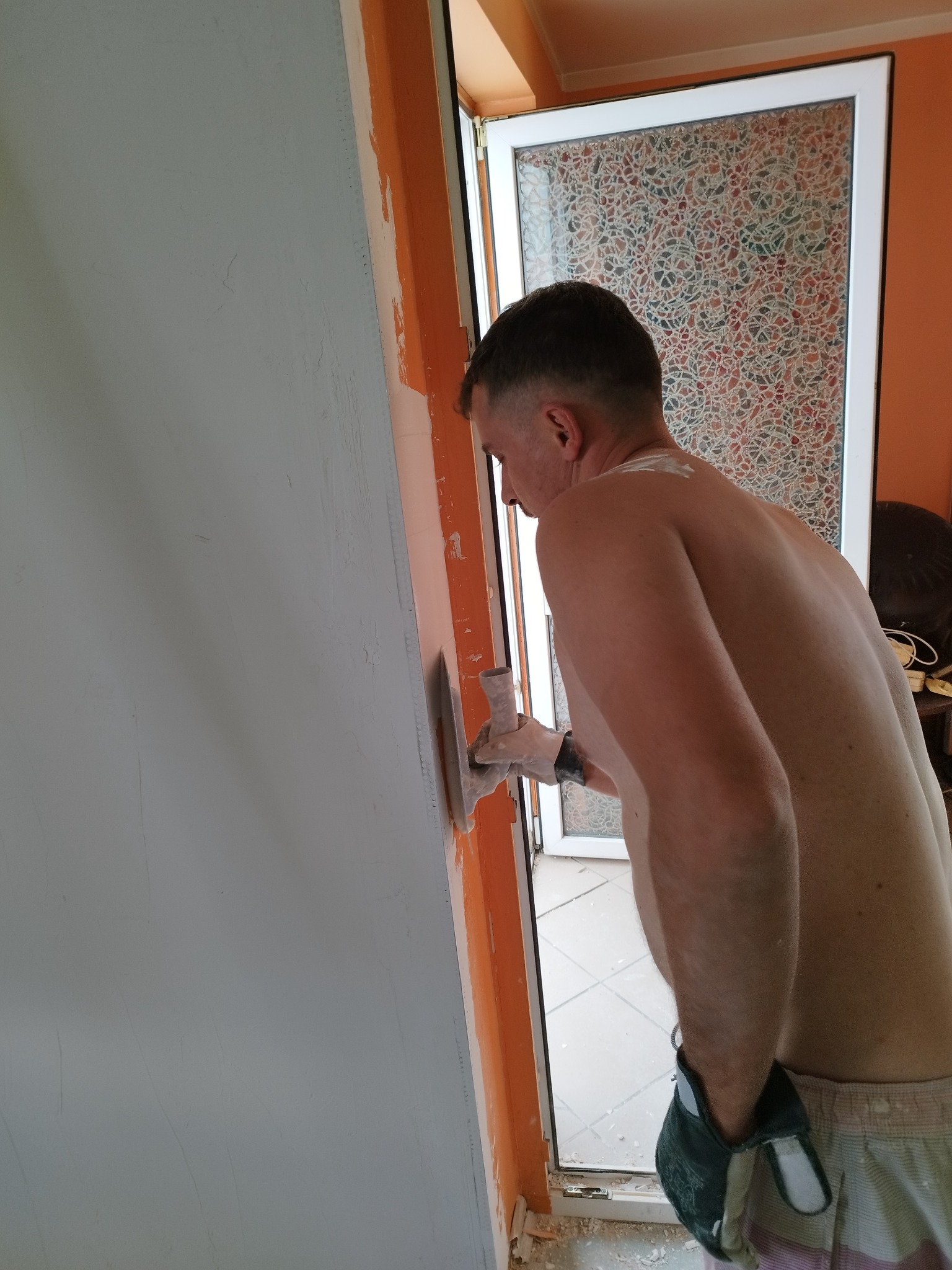 Mężczyzna bez koszulki, w rękawicach, wygładza szpachlą ścianę w trakcie remontu, widoczne niedoskonałości na powierzchni.