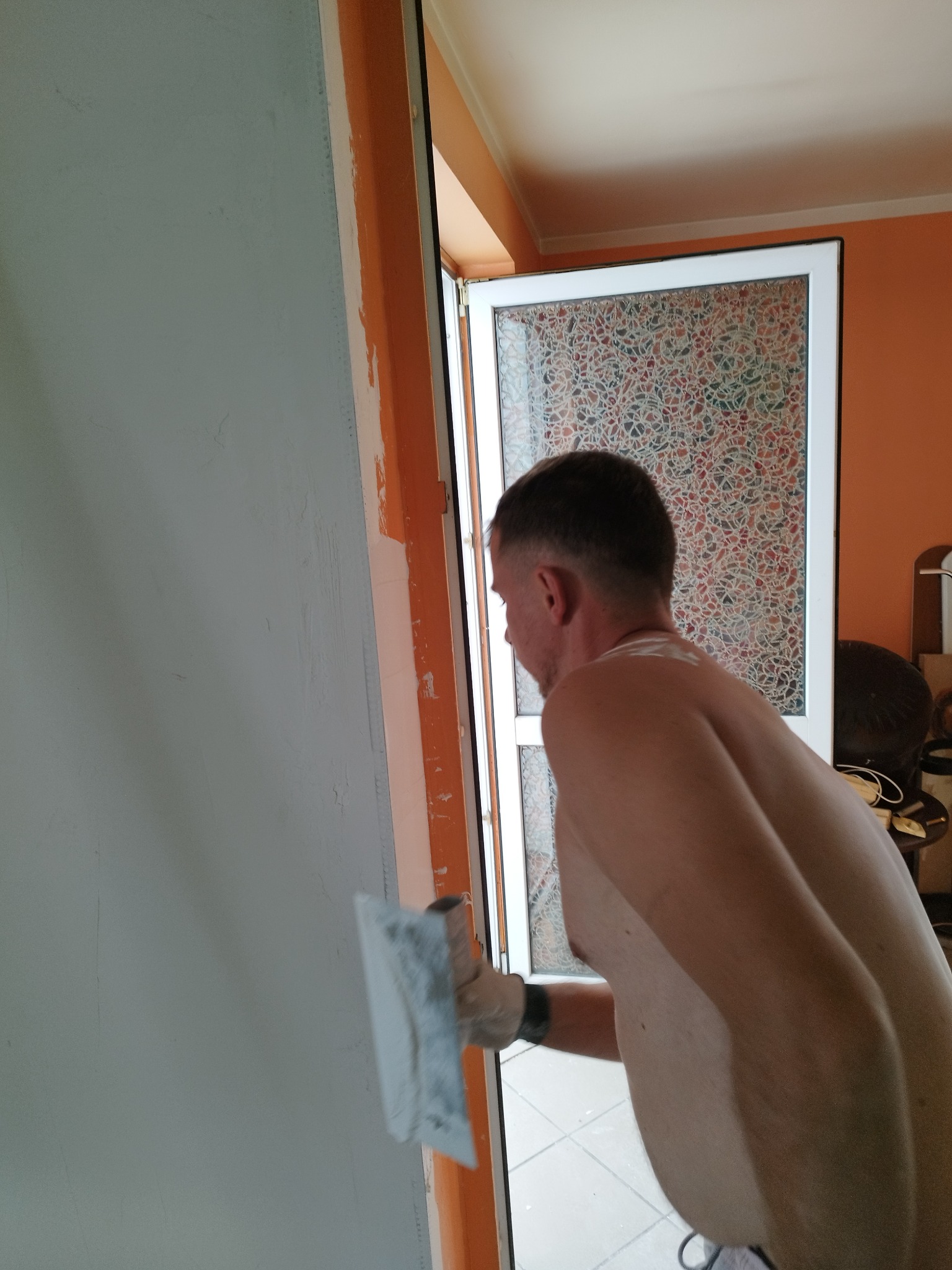 Mężczyzna w rękawicach wyrównuje szpachelką ścianę wewnątrz pomieszczenia, widoczna pomarańczowa futryna drzwi i wzorzysta szyba.