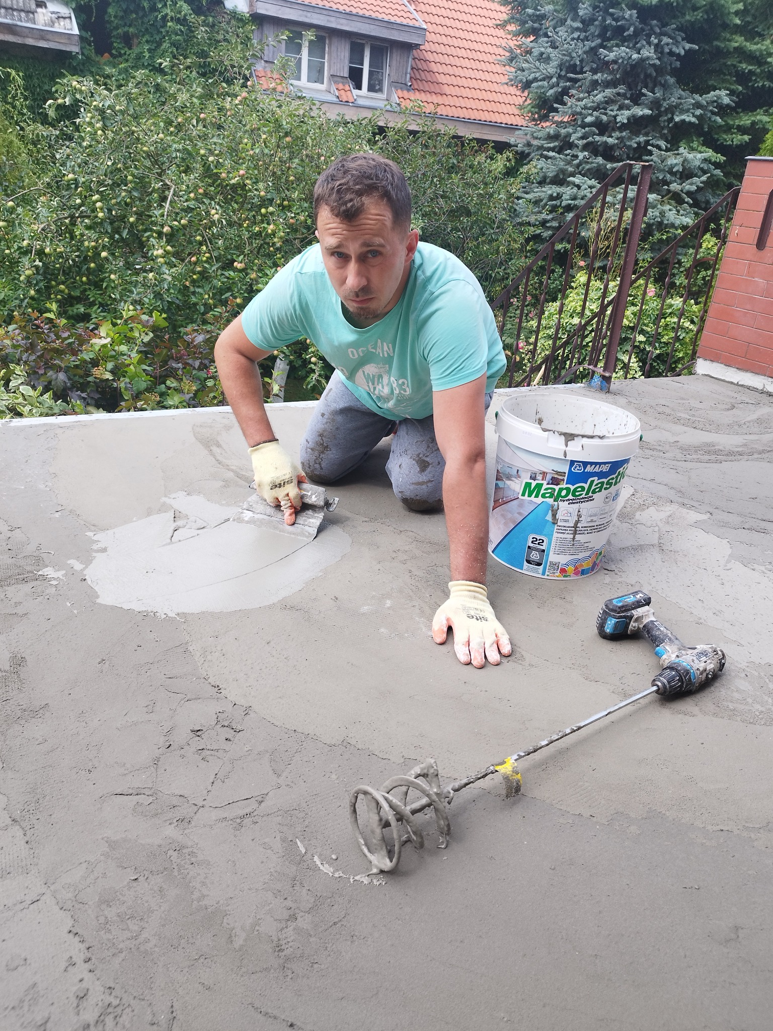 Pracownik w rękawicach wyrównuje szpachelką warstwę hydroizolacji na powierzchni, obok stoi wiadro z masą Mapelastic i mieszadło elektryczne.
