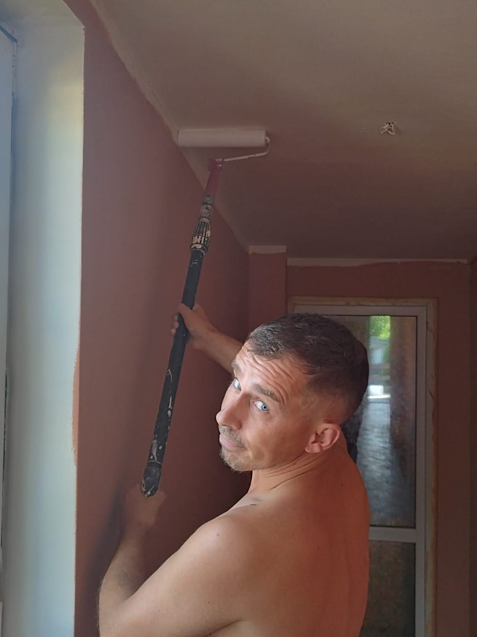 Mężczyzna bez koszulki maluje sufit wałkiem na przedłużonym kiju, spoglądając w bok z uśmiechem.