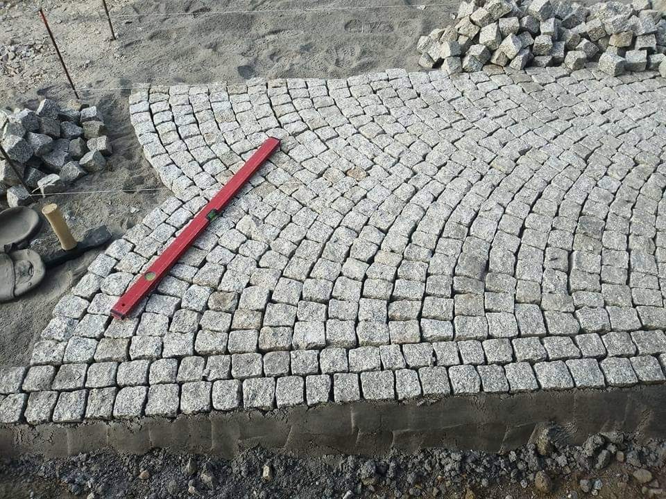 Układanie kostki z granitu w łukowaty wzór, widoczna poziomica, zapas kostki obok i fragment podsypki piaskowej.