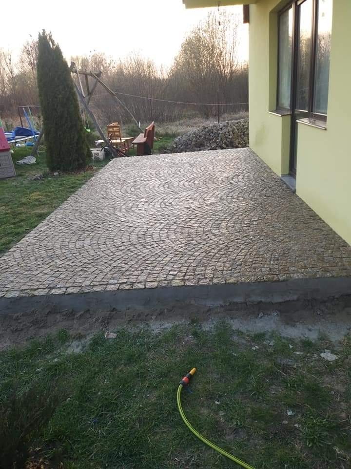 Nowa nawierzchnia z kostki granitowej ułożona w półokręgi, przylegająca do budynku z żółtą elewacją; widoczny wąż ogrodowy na trawie.