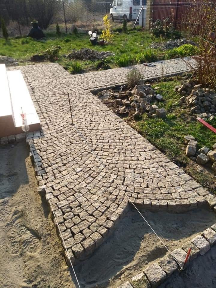 Kładzenie ścieżki z kostki granitowej w ogrodzie, widoczne sznurki wyznaczające granice, etap prac ziemnych i częściowo ułożona nawierzchnia w charakterystyczny łukowy wzór.