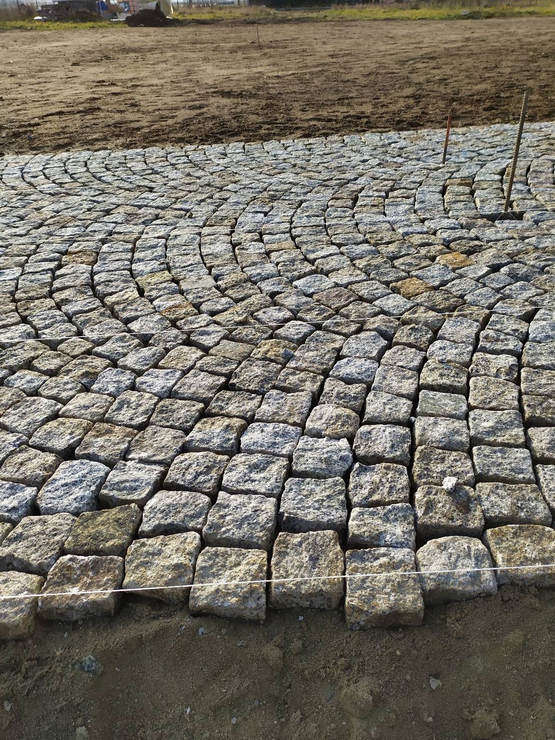 Fragment układanej nawierzchni z kostki granitowej w kształcie łuku, widoczne sznurki wyznaczające linię, ziemia w tle.