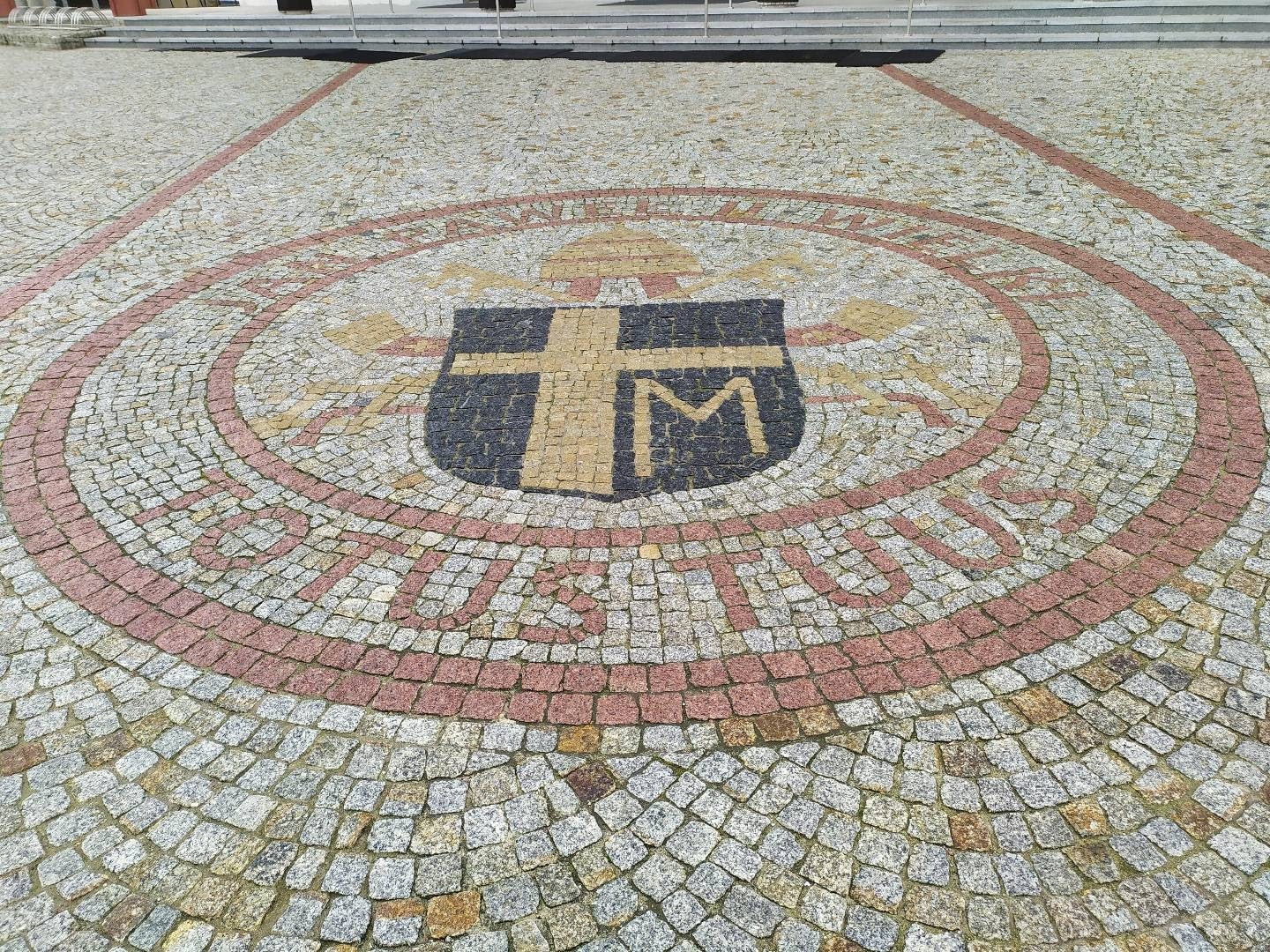 Granitowa kostka brukowa ułożona w okrągły wzór herbu z napisem 'TOTUS TUUS' na placu przed budynkiem.