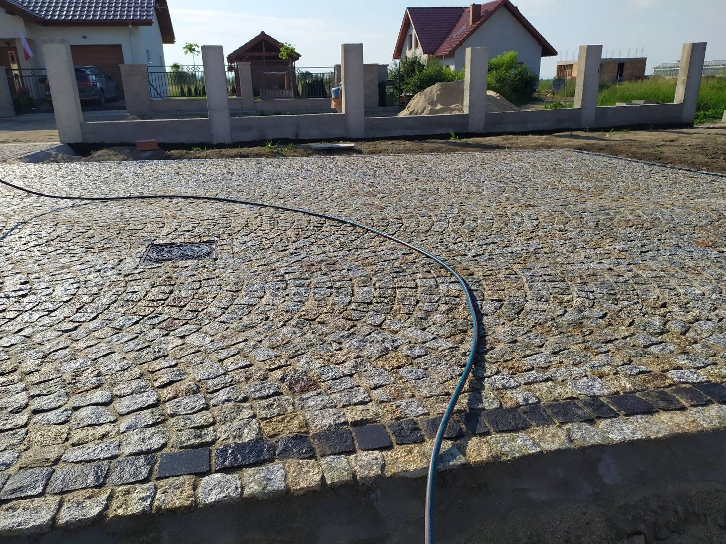 Ułożona kostka granitowa w radialny wzór z ciemniejszym obramowaniem, widoczny właz kanalizacyjny i niebieski wąż ogrodowy, w tle domy w trakcie budowy.