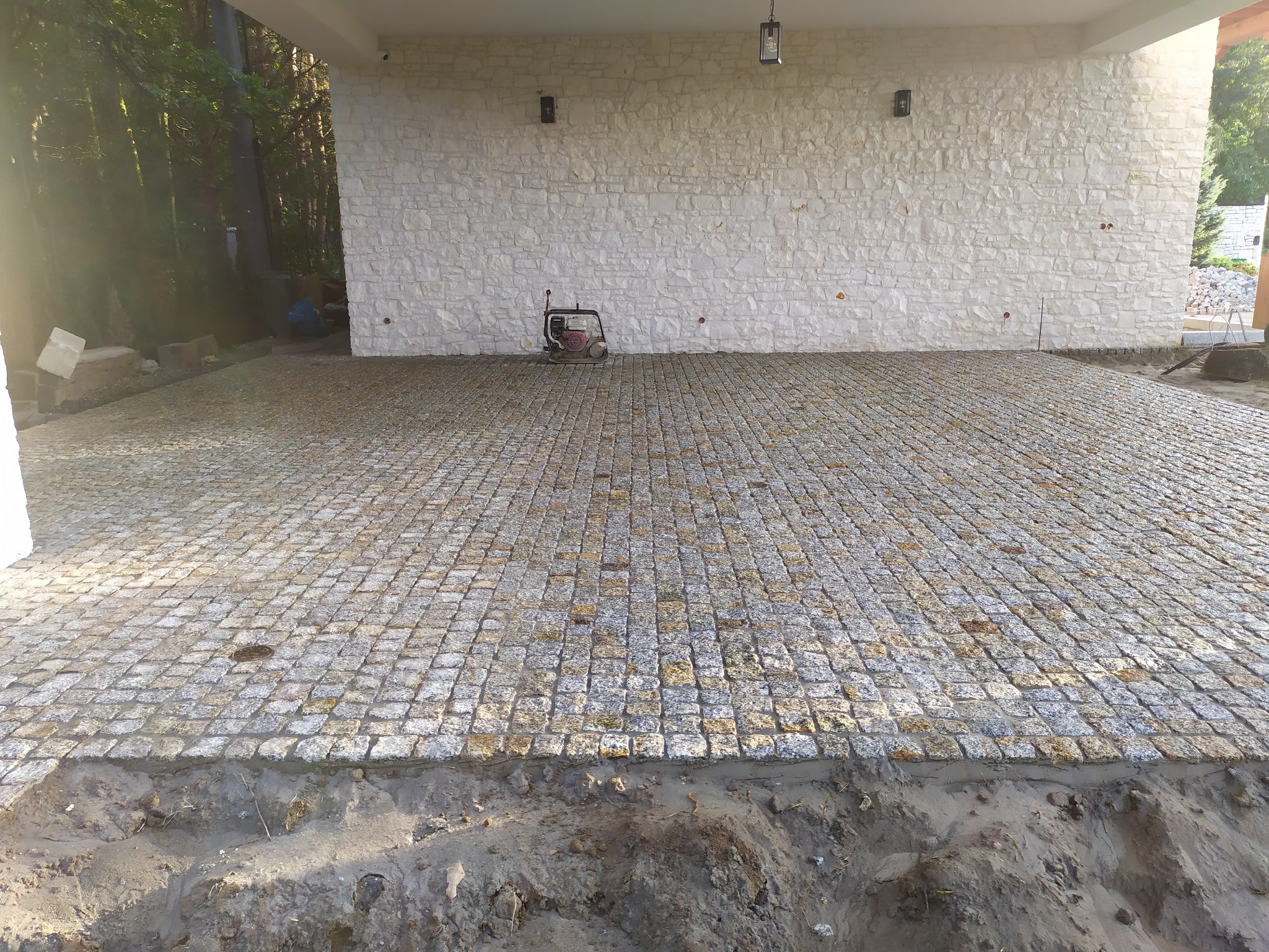 Nowa nawierzchnia z kostki granitowej przed budynkiem z elewacją z kamienia naturalnego; widoczny zagęszczarka wibracyjna.
