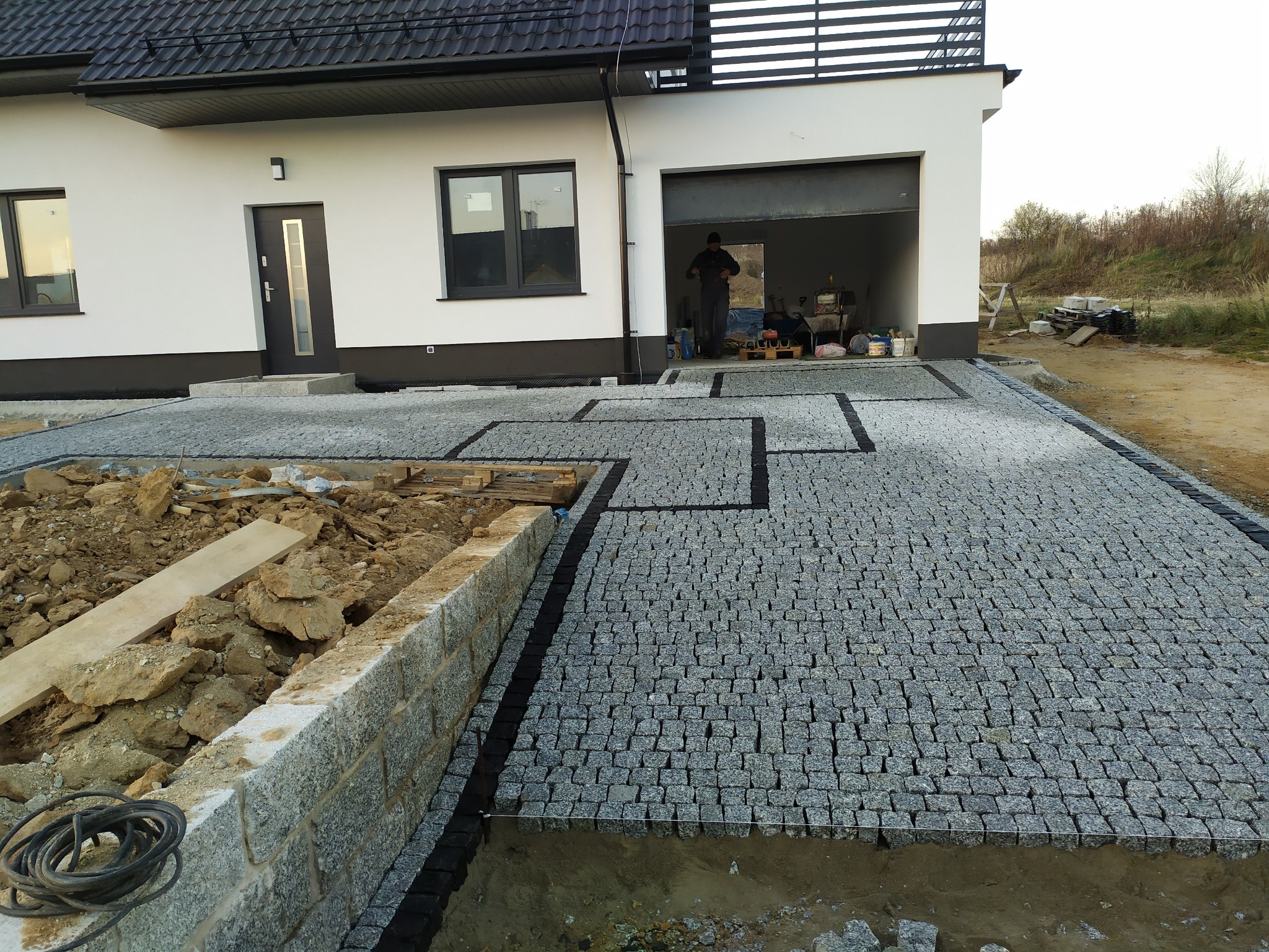 Częściowo ułożona szara kostka na podjeździe przed nowoczesnym domem z garażem, widoczny pracownik w garażu i przygotowany teren pod dalsze prace brukarskie.