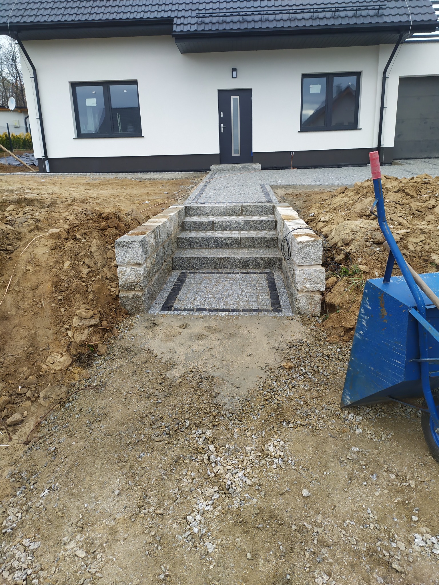 Granitowe schody prowadzące do nowoczesnego domu jednorodzinnego, widoczne prace ziemne i niebieska taczka.