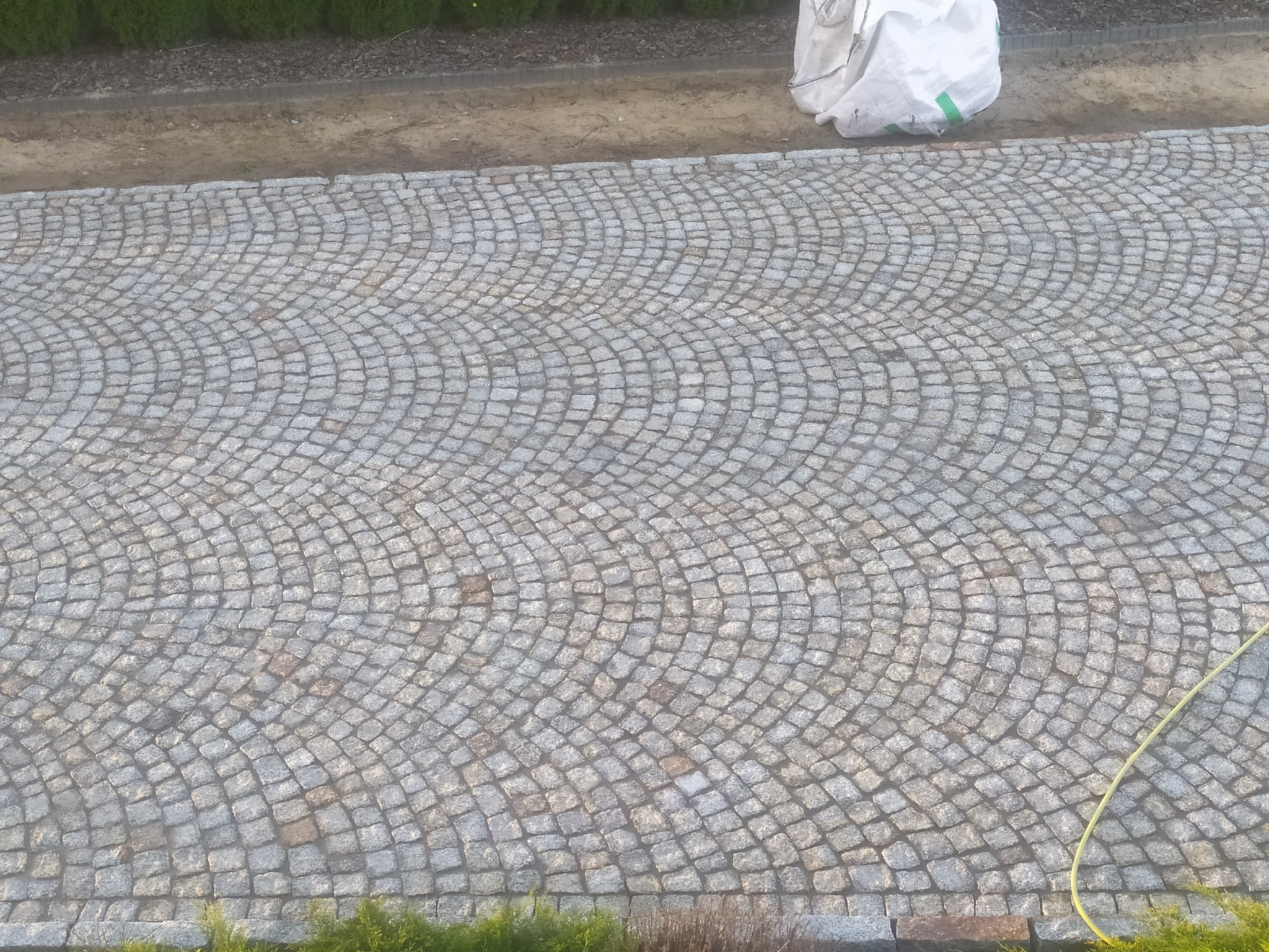 Szara kostka granitowa ułożona w łukowaty wzór, widoczny fragment węża ogrodowego i worka z materiałem budowlanym w tle.