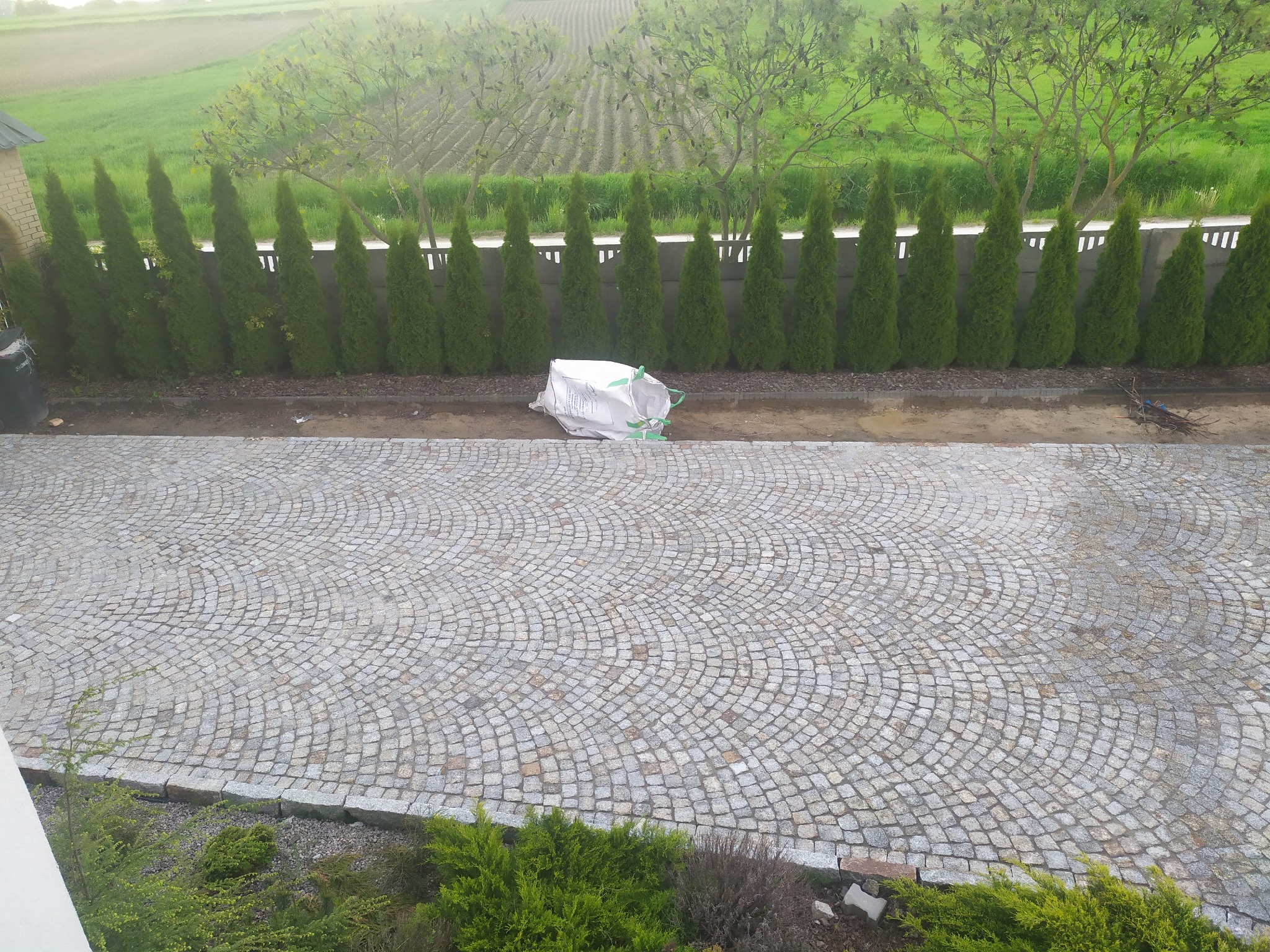Ułożona kostka granitowa w okrągły wzór na podjeździe, z workiem materiału budowlanego w tle i rzędem tuj wzdłuż ogrodzenia.