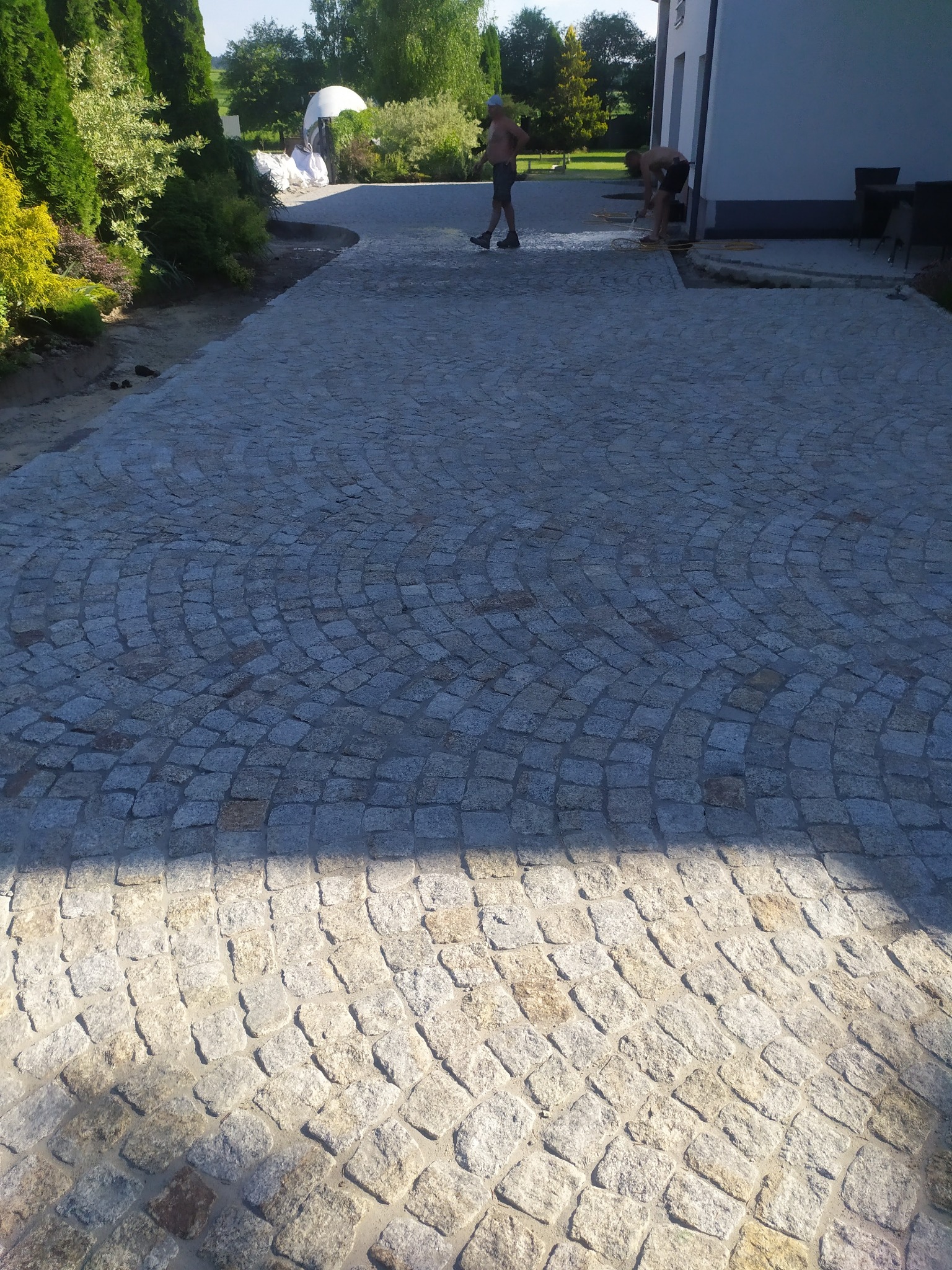 Ułożona nawierzchnia z kostki granitowej w kształcie wachlarza, z pracownikami w tle na posesji domu jednorodzinnego.
