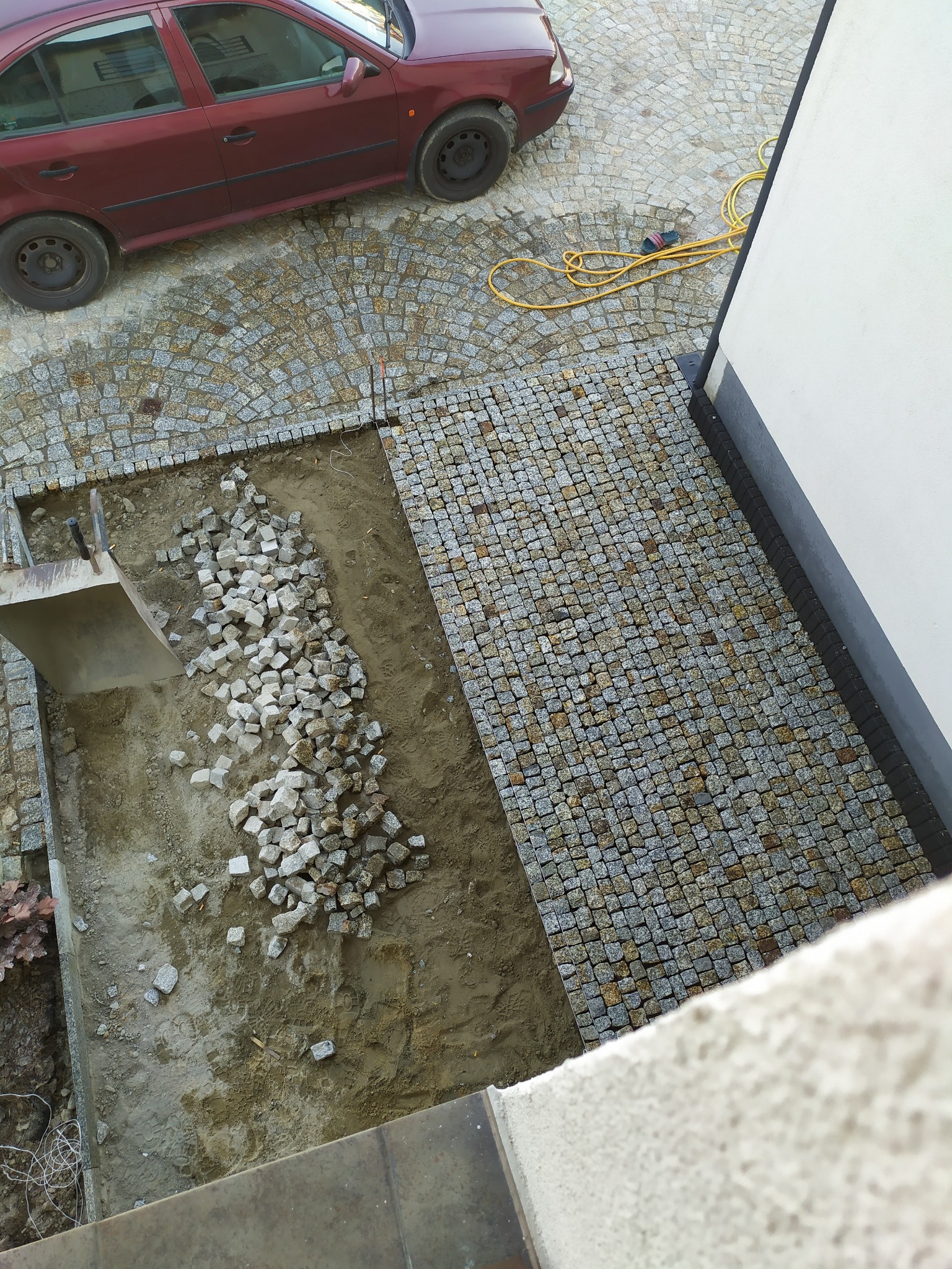 Widok z góry na częściowo ułożoną kostkę brukową, z hałdą kamieni i taczką w przygotowanym wykopie, obok zaparkowany bordowy samochód i zwinięty żółty przedłużacz.