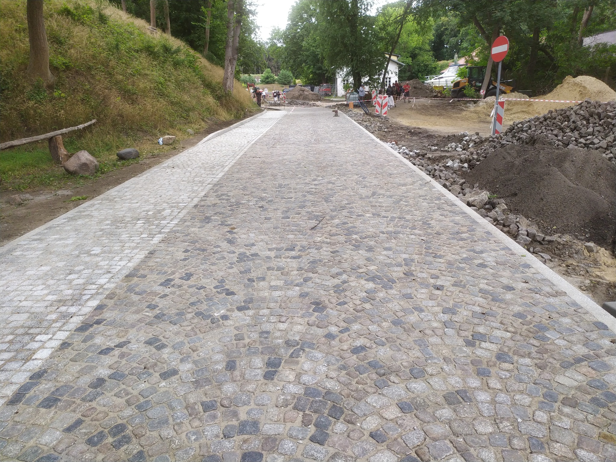 Nowa ścieżka z kostki granitowej ułożona w łukowaty wzór, z fragmentem wykończonym jaśniejszą kostką, w tle widoczne prace ziemne i robotnicy.