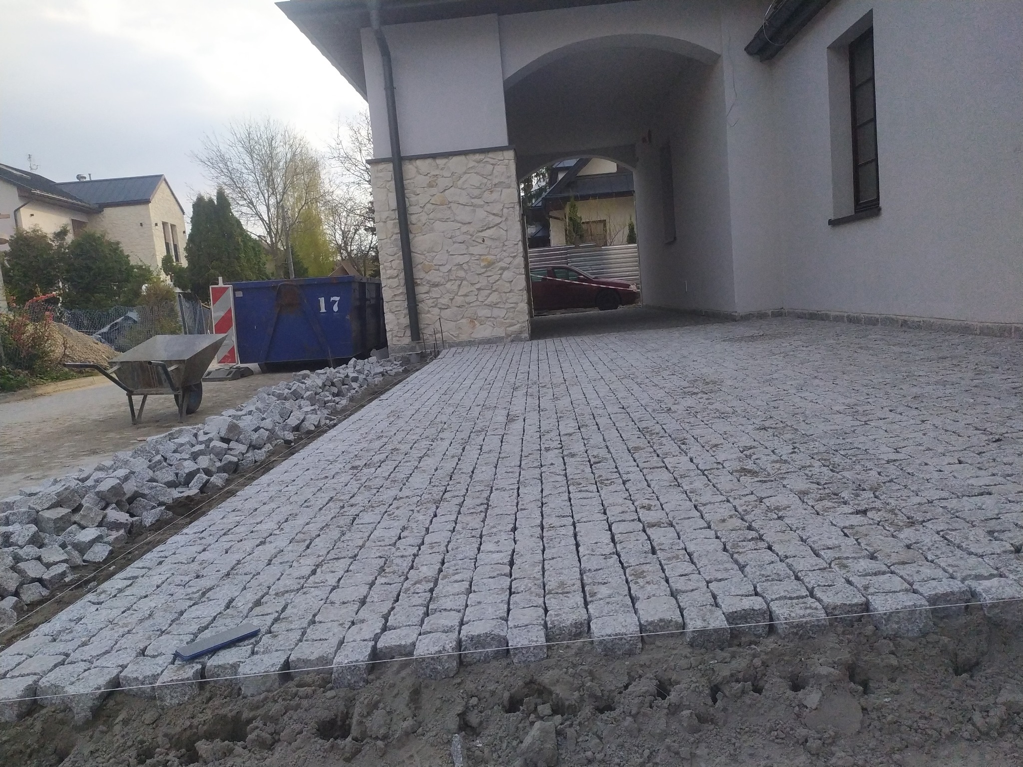 Układanie szarej kostki granitowej na podjeździe, widoczny fragment ułożonej nawierzchni, przygotowane kamienie, taczka i kontener budowlany w tle, prace w toku.