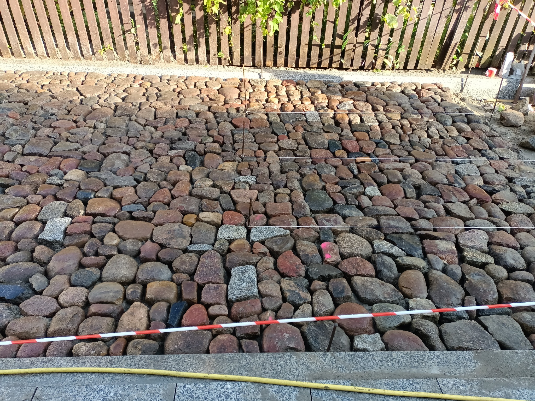 Nierówna powierzchnia z ułożonych, różnokolorowych kamieni polnych, oddzielona taśmą ostrzegawczą od chodnika, z drewnianym płotem w tle i zielenią zwisającą z góry.