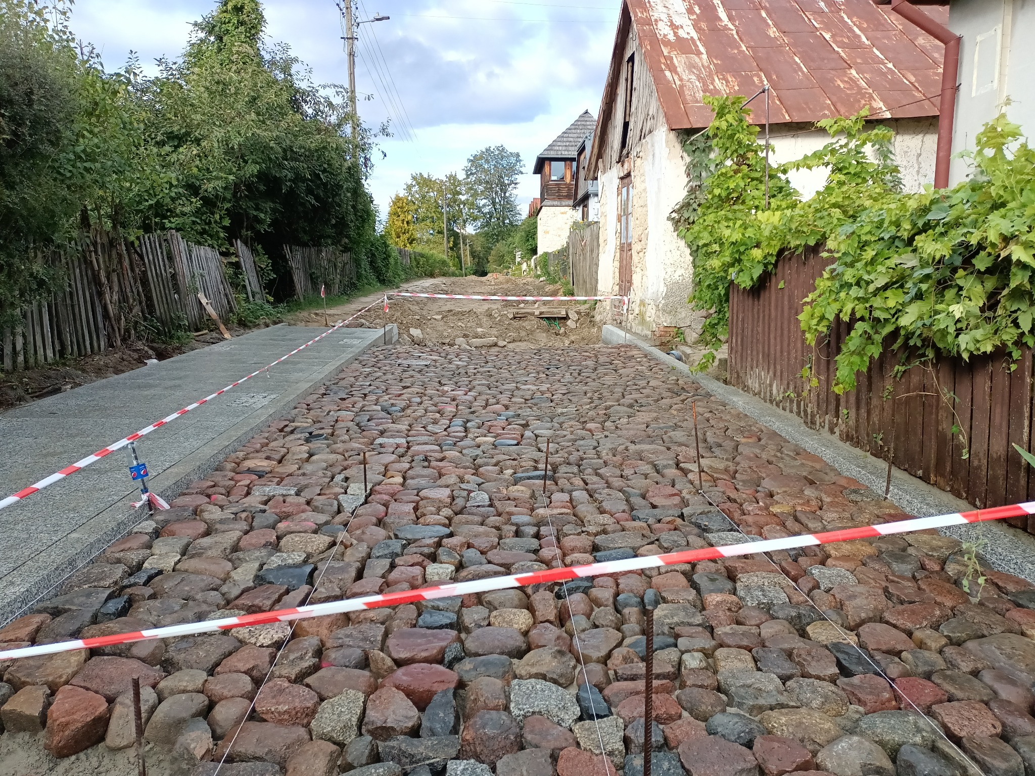 Ulica w trakcie układania z kostki brukowej, zabezpieczona taśmą ostrzegawczą, z widocznymi budynkami w tle i fragmentami odsłoniętej ziemi.