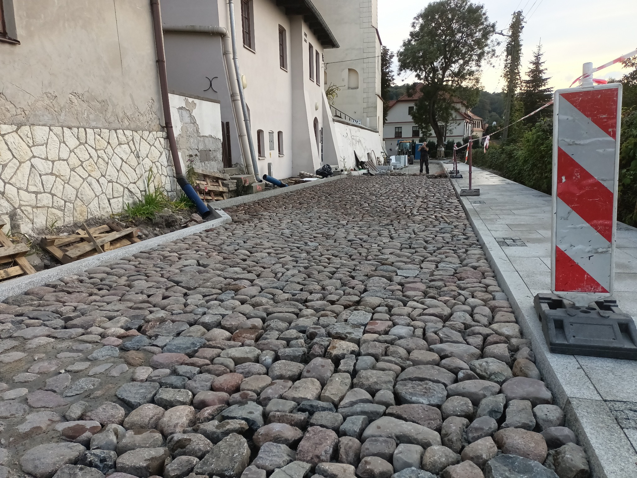 Ułożona nawierzchnia z nieregularnych kamieni na ulicy w pobliżu budynku, z widocznymi pracownikami w tle i znakiem drogowym po prawej stronie.