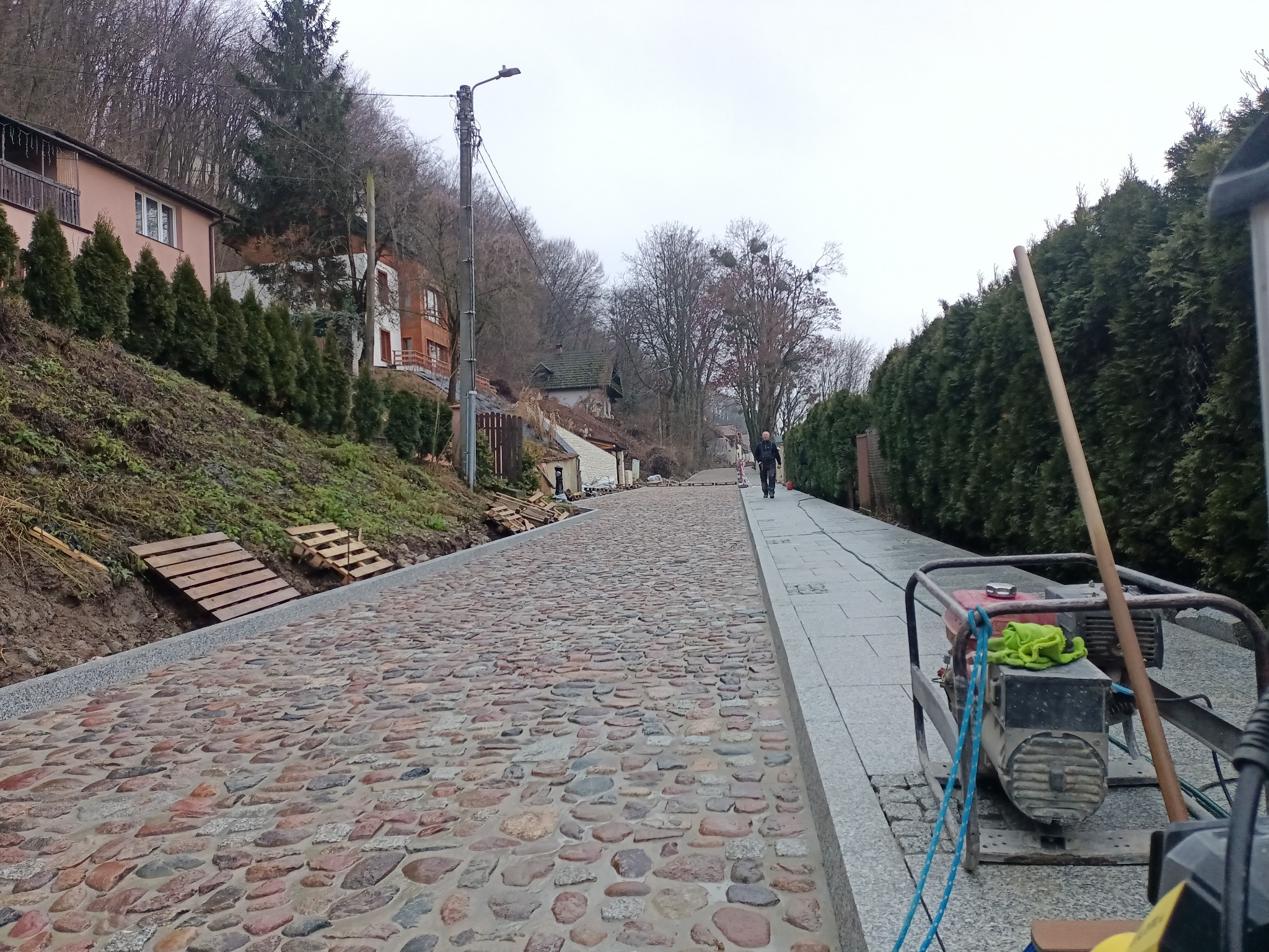 Ulica wyłożona czerwonobrązową kostką brukową, po prawej stronie chodnik z jasnych płyt, w tle budynki mieszkalne na wzgórzu, widoczny generator prądu i drewniane pomosty na zboczu.