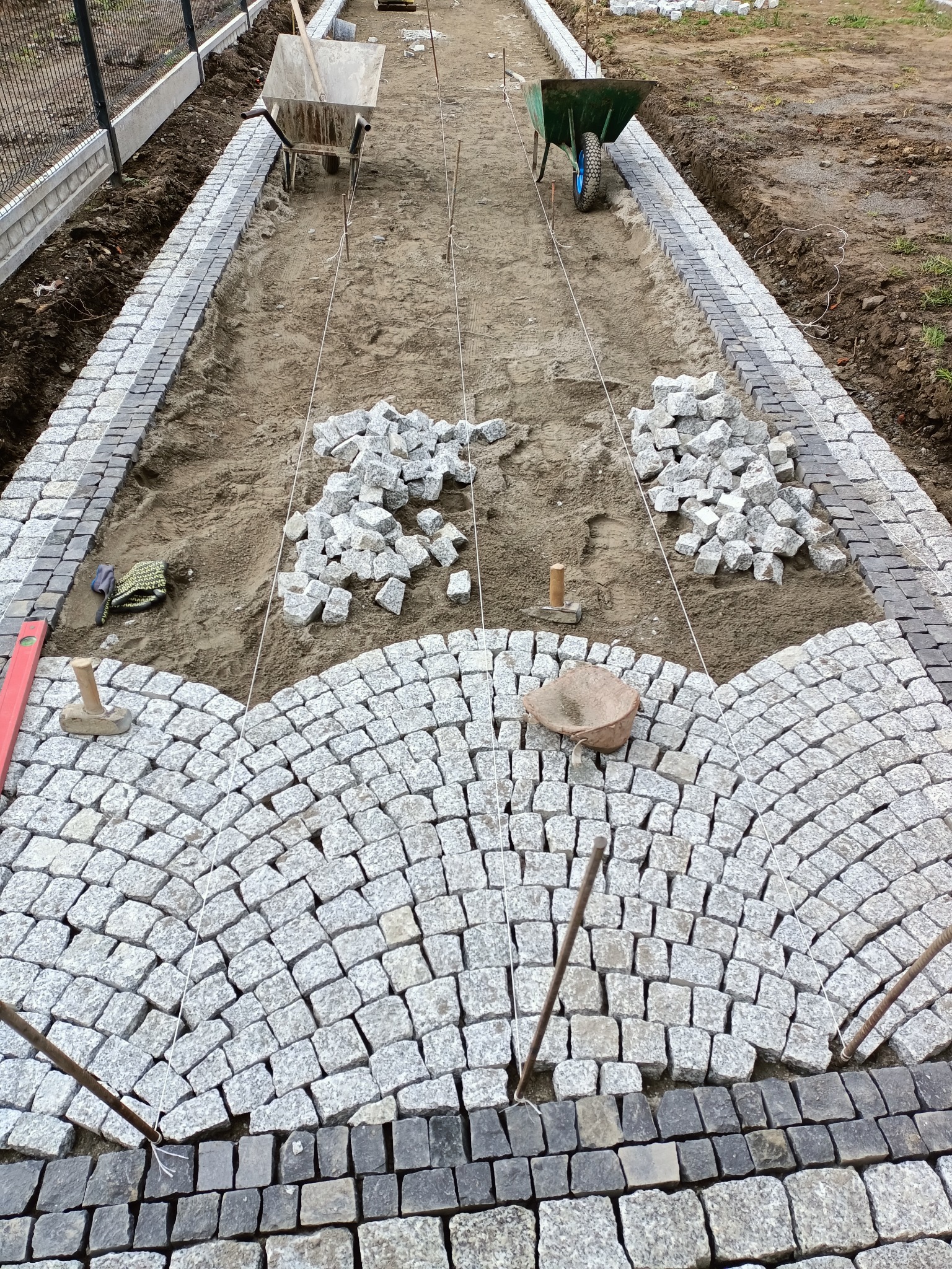 Widok z góry na proces układania chodnika z kostki granitowej w łukowaty wzór, z widocznymi narzędziami, takimi jak poziomica, młotek brukarski, sznurki wyznaczające linię, oraz dwie taczki w tle.