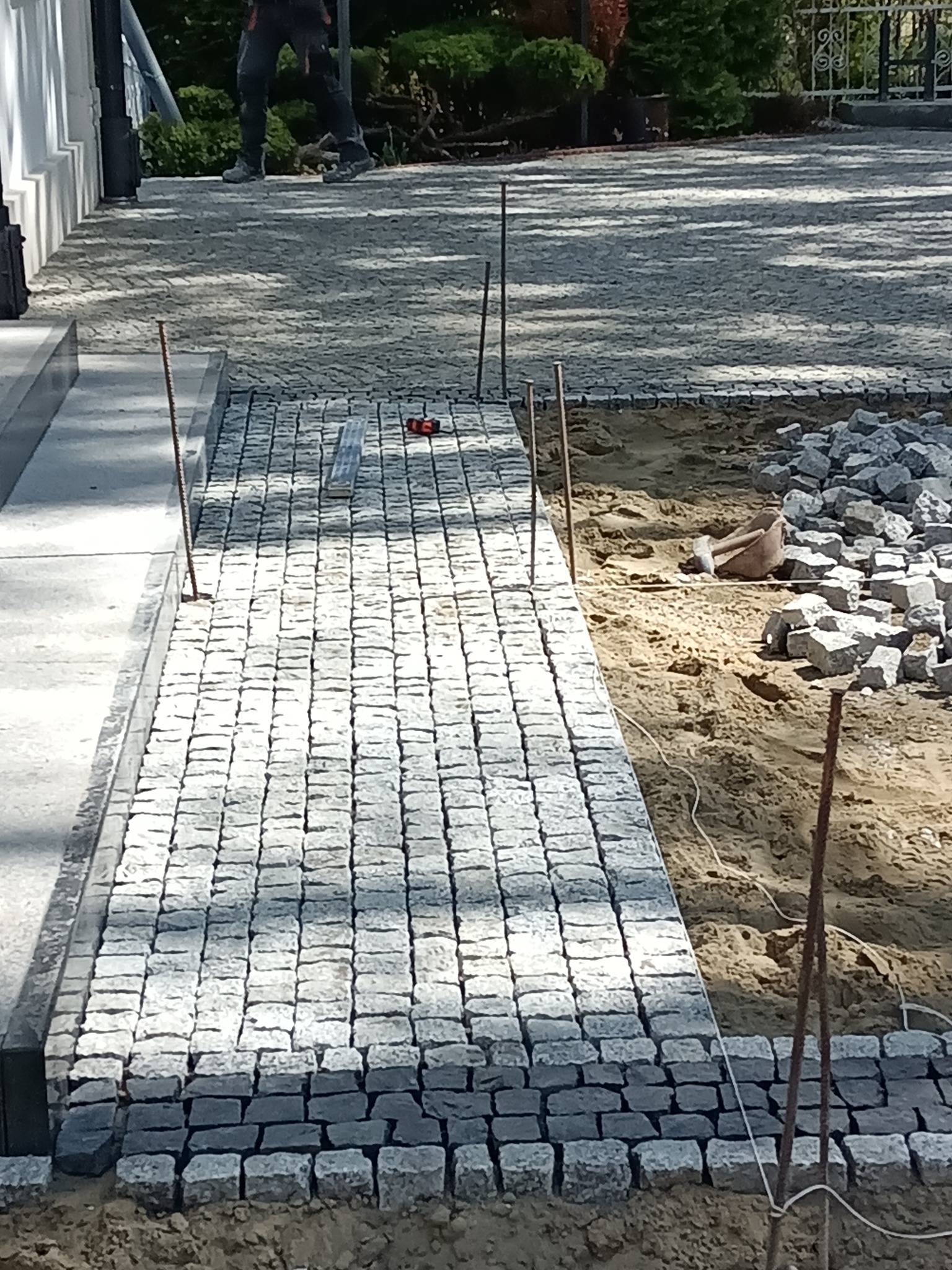 Układanie kostki granitowej w trakcie prac: widoczne fragmenty ułożonej nawierzchni, przygotowane podłoże z piasku, stos kostki brukowej i narzędzia budowlane, całość w otoczeniu zieleni...