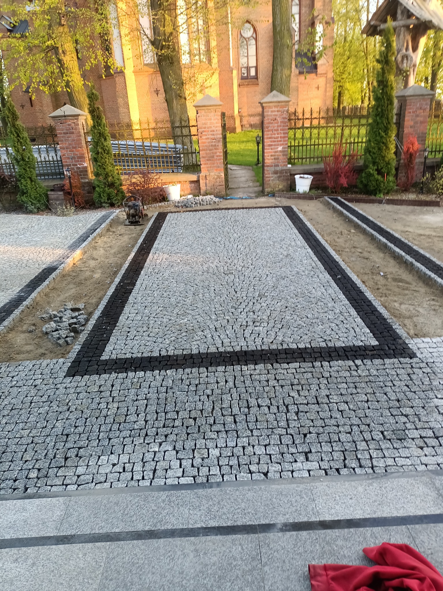 Fragment placu z szarej kostki granitowej z czarną obwódką, w tle budynek z cegły i zieleń, widoczna ubijarka do ziemi i przygotowane podłoże pod kolejne fragmenty nawierzchni.