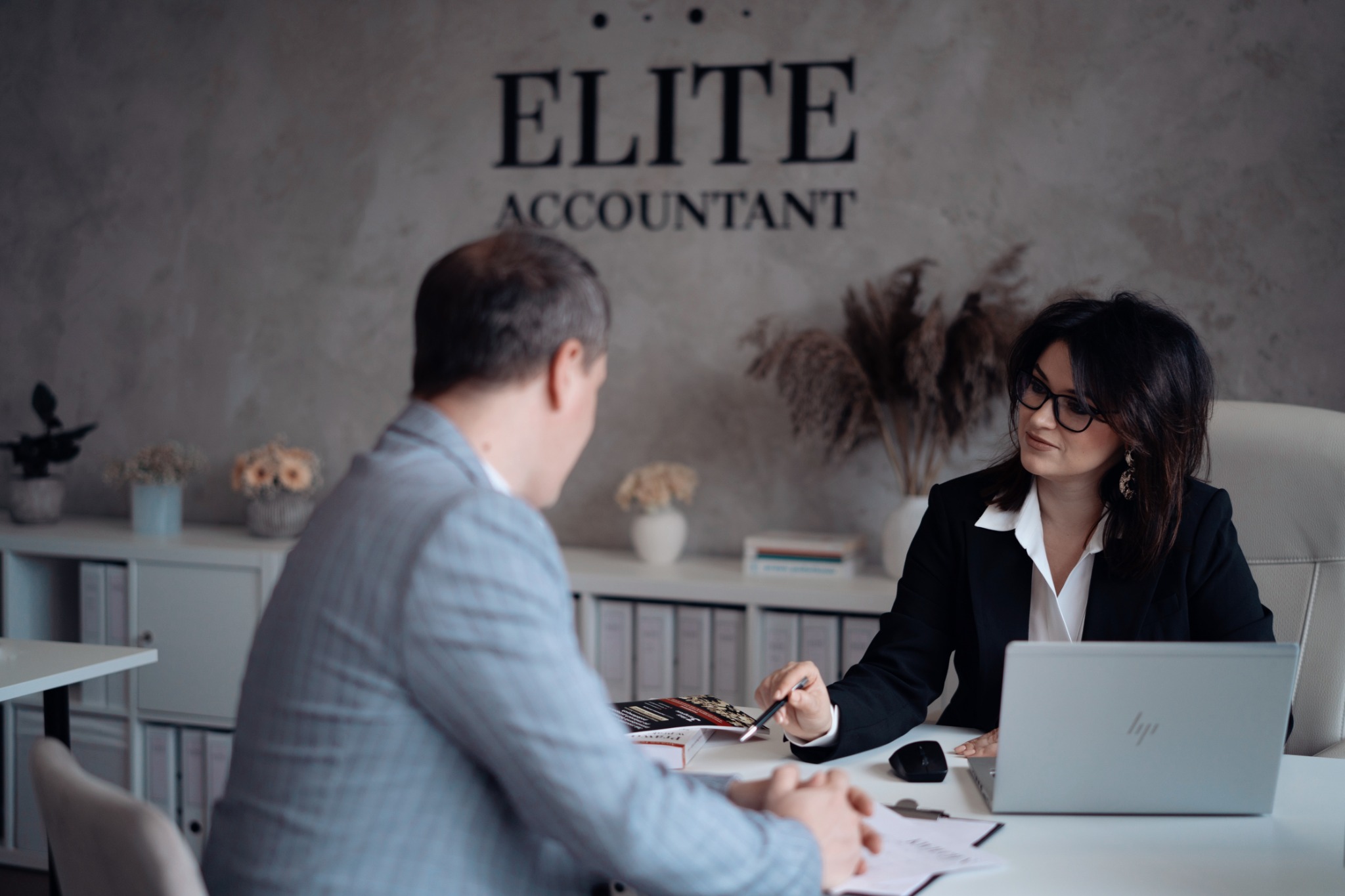 Konsultacja w biurze: kobieta w okularach i marynarce omawia dokumenty z klientem, w tle logo 'Elite Accountant' i dekoracje.