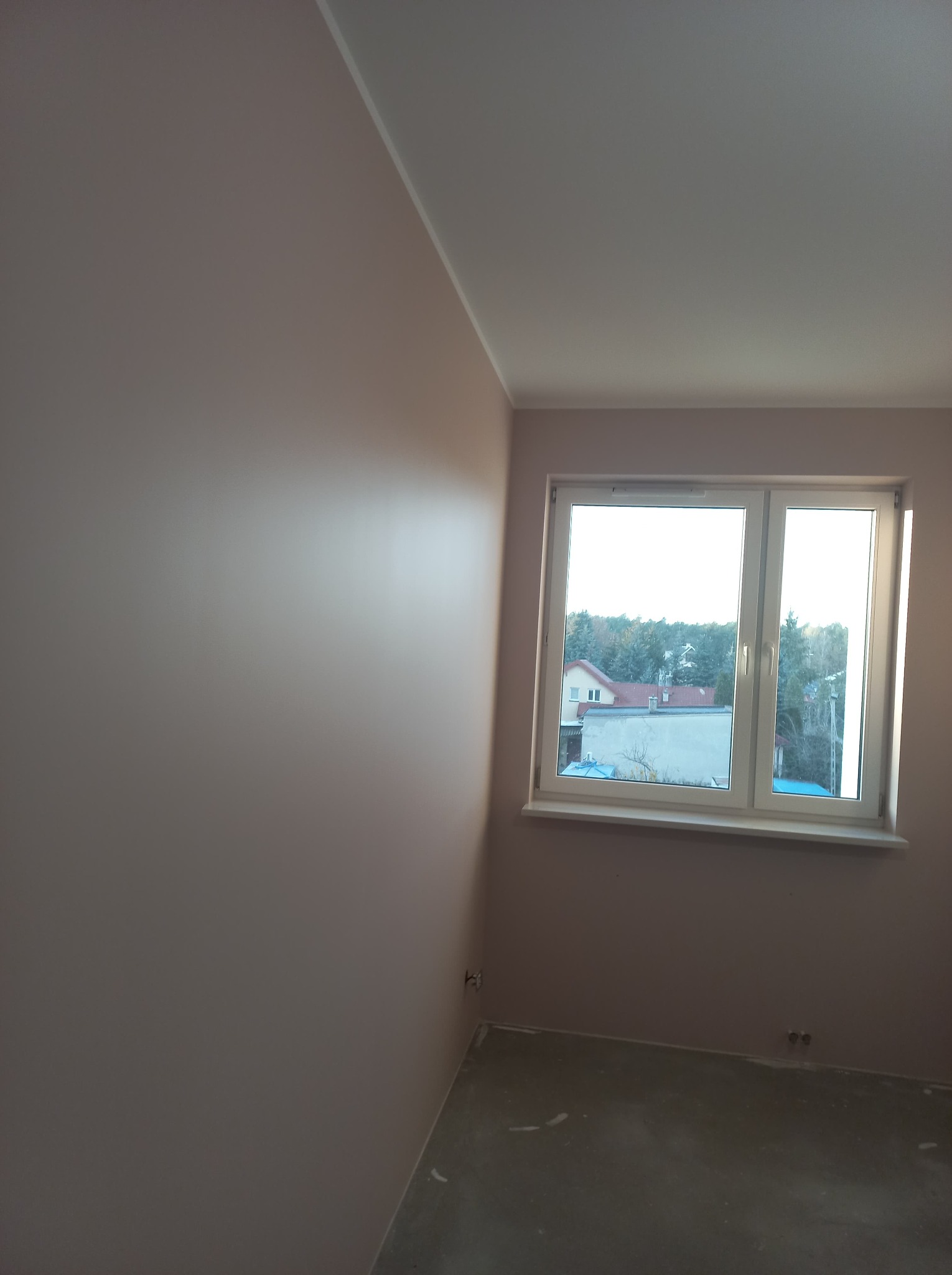 Pomalowane ściany w pokoju na delikatny, pudrowy róż, widok na okno z widokiem na drzewa i budynki w tle, surowa podłoga w trakcie prac wykończeniowych.