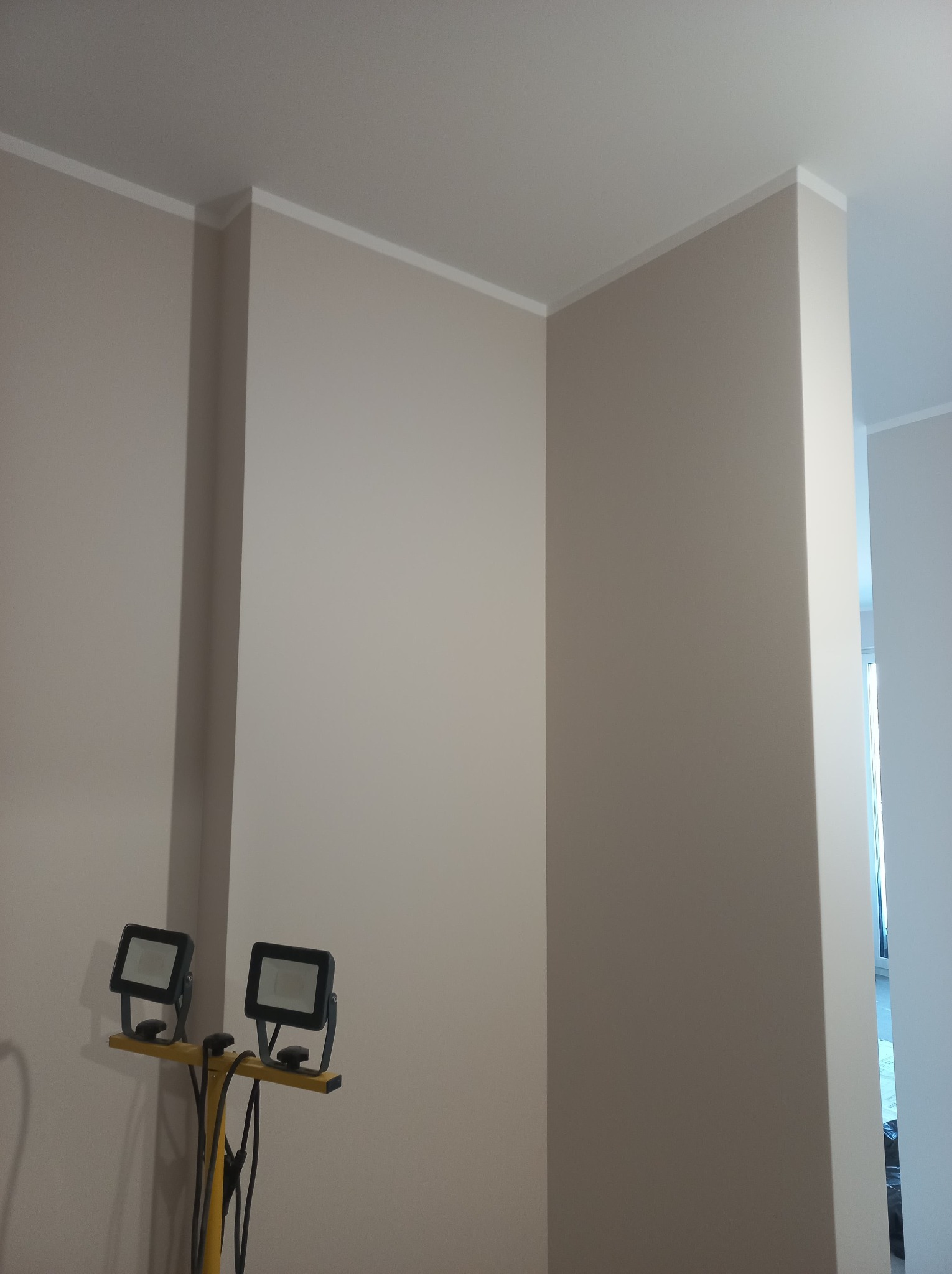 Świeżo pomalowane ściany w odcieniach beżu z białym wykończeniem sufitu, oświetlone dwoma halogenami na żółtym statywie.