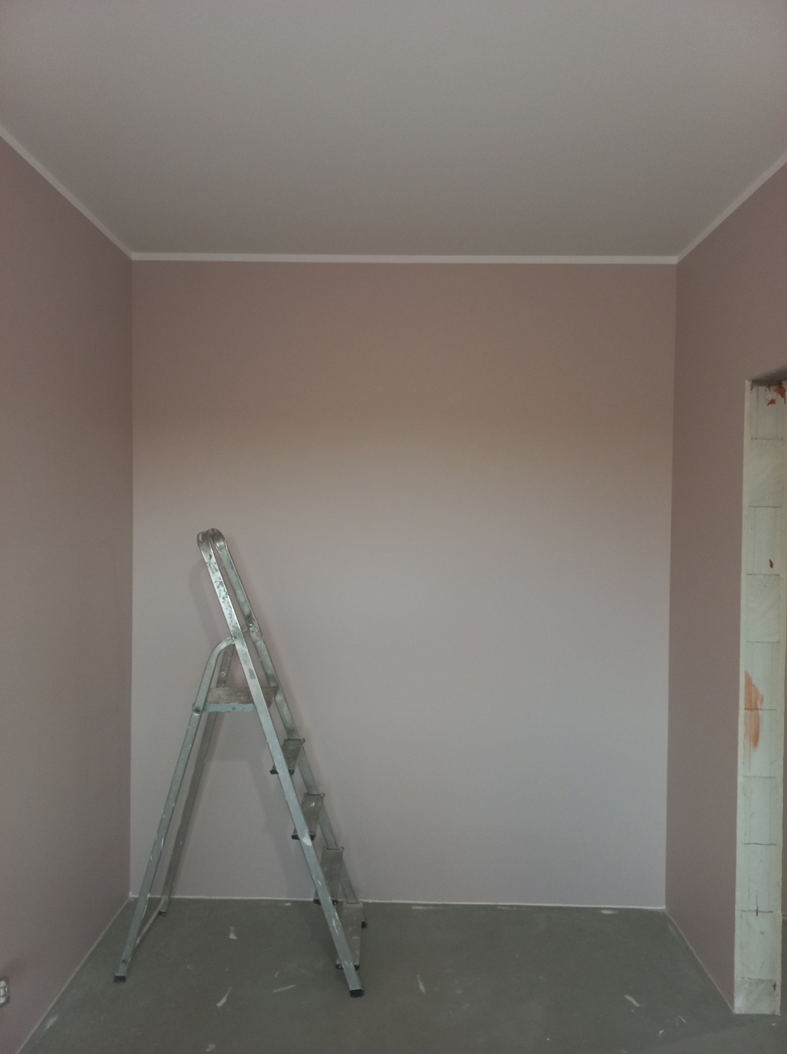 Pomieszczenie po malowaniu ścian na delikatny róż, z widoczną aluminiową drabiną i fragmentem surowego muru w przejściu.