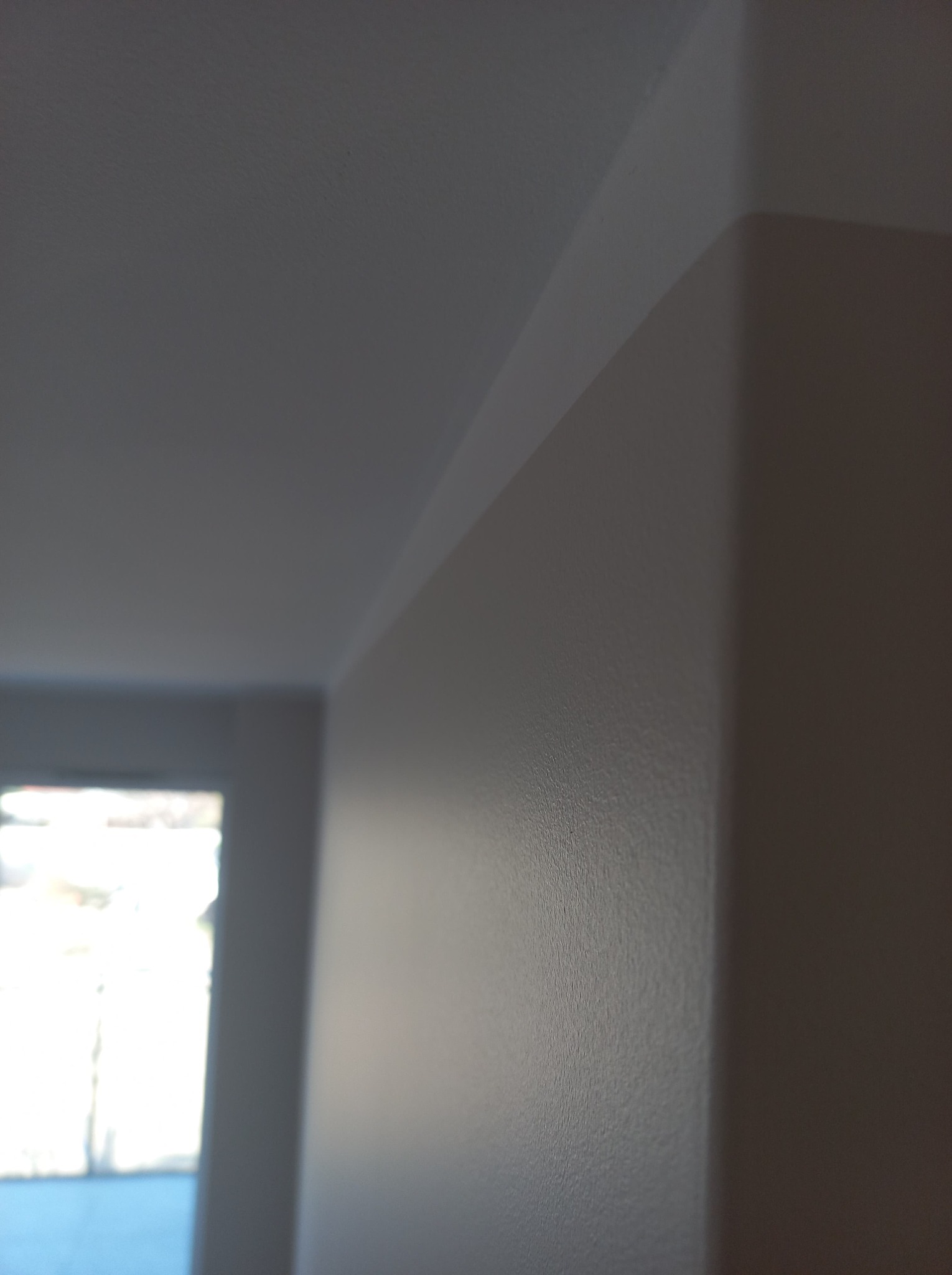 Pomalowany narożnik pokoju w odcieniach beżu i bieli, z widoczną fakturą ściany i fragmentem okna w tle.