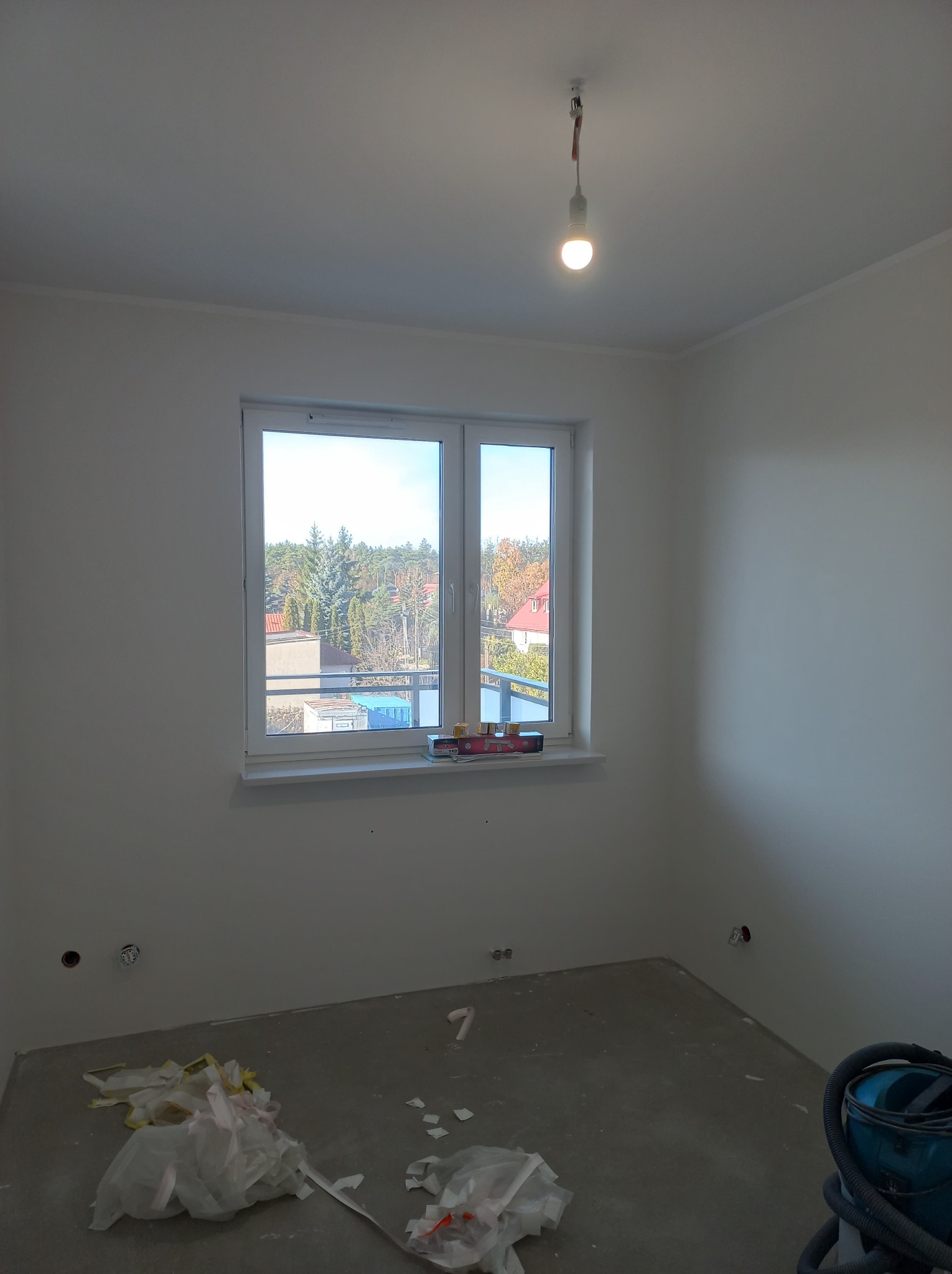 Pokój po szpachlowaniu i wstępnym malowaniu na biało. Widoczne ślady po gładzi na podłodze, narzędzia na parapecie i wystające kable z sufitu.