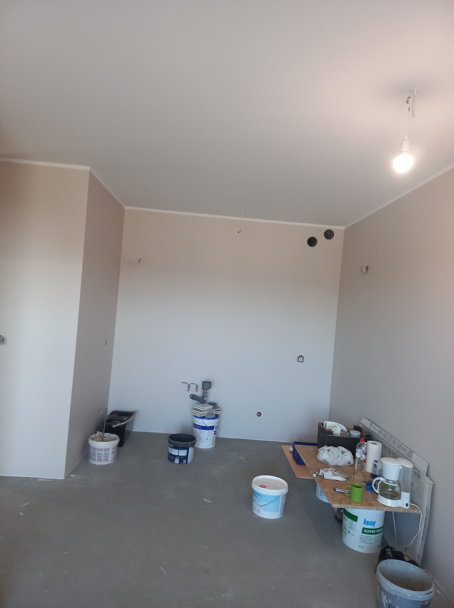 Wnętrze pokoju w trakcie prac wykończeniowych; ściany pomalowane na jasny beż, widoczne puszki z farbą, narzędzia malarskie i stolik z akcesoriami. Na ścianie widoczne punkty instalacji elektrycznej.