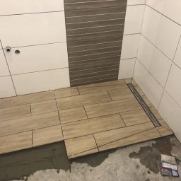 REMHOME - Układanie beżowych płytek podłogowych w łazience z odpływem liniowym; widoczne krzyżyki dystansowe i zaprawa klejowa.