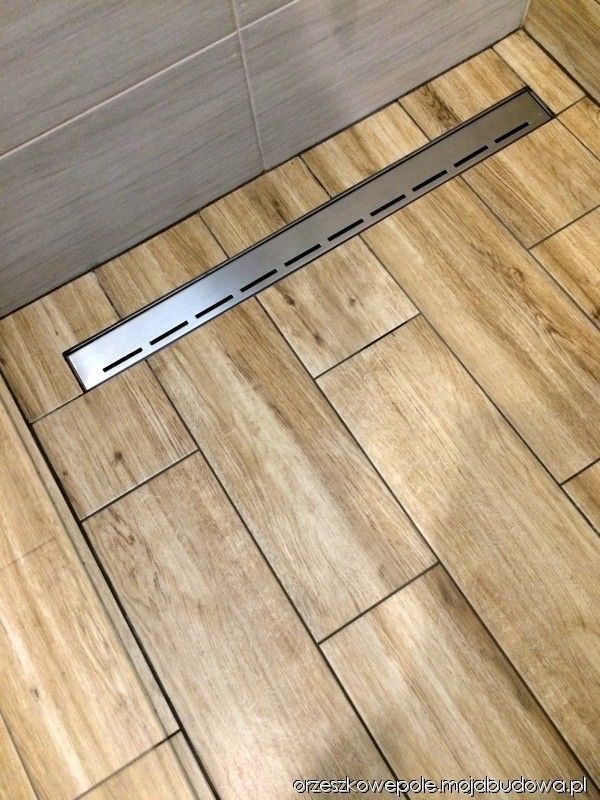 Ułożona pod kątem podłoga z płytek imitujących drewno w łazience z zamontowanym liniowym odpływem prysznicowym ze stali nierdzewnej z podłużnymi szczelinami.