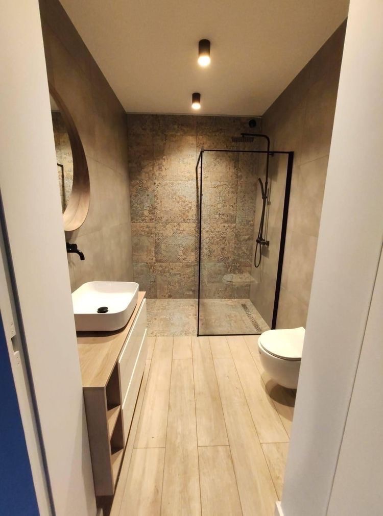 Nowoczesna łazienka z kabiną prysznicową typu walk-in z czarną ramą, umywalką nablatową na szafce z drewnianym blatem, okrągłym lustrem i płytkami w odcieniach beżu i brązu.
