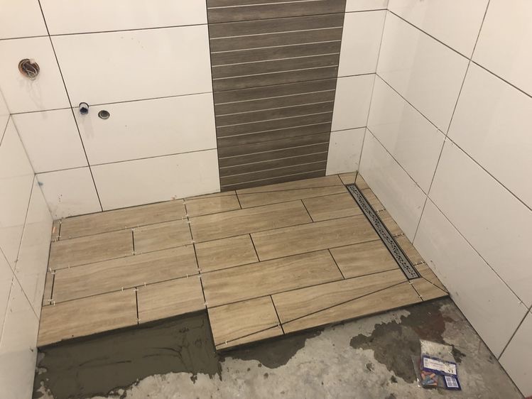 Układanie beżowych płytek podłogowych w łazience z odpływem liniowym; widoczne krzyżyki dystansowe i zaprawa klejowa.
