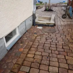 Ułożona z czerwono-brązowej kostki granitowej ścieżka obok budynku z widocznym oknem piwnicznym, w tle jezioro i wózek budowlany.