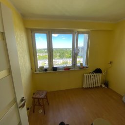 Mateusz Warot - Pokój z żółtymi ścianami przed remontem: widoczne stare tapety, grzejnik, okno z widokiem na zieleń. Na podłodze leżą narzędzia i widać kota. Przygotowanie do malowania.