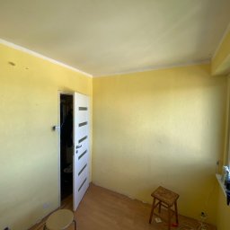 Mateusz Warot - Żółty pokój przed remontem: ściany z widocznymi niedoskonałościami, otwarte białe drzwi, dwa stołki i okno z akcesoriami malarskimi na parapecie. Widok z góry.