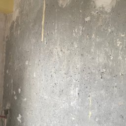 Mateusz Warot - Szara, surowa ściana z widocznymi śladami po usuniętych tapetach i resztkami starej farby, przygotowana do dalszych prac wykończeniowych. Fragment żółtej ściany w rogu.