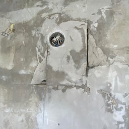 Mateusz Warot - Ściana w trakcie remontu z widocznym otworem na puszkę elektryczną i fragmentami instalacji. Widoczne ślady po szpachlowaniu i nierówności na powierzchni.