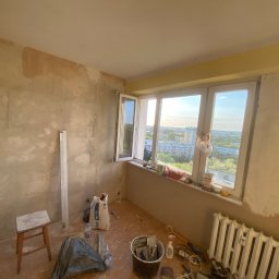 Mateusz Warot - Pokój w trakcie szpachlowania: ściany częściowo pokryte gładzią, widoczne narzędzia, worek z gipsem MP 75, okno z widokiem na miasto, grzejnik i krzesło.