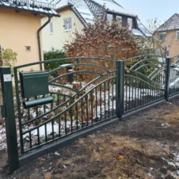 Nowoczesne, ciemnozielone ogrodzenie z metaloplastyki z przymocowaną skrzynką na listy, ustawione na świeżo przekopanym terenie z resztkami śniegu, na tle domów jednorodzinnych.