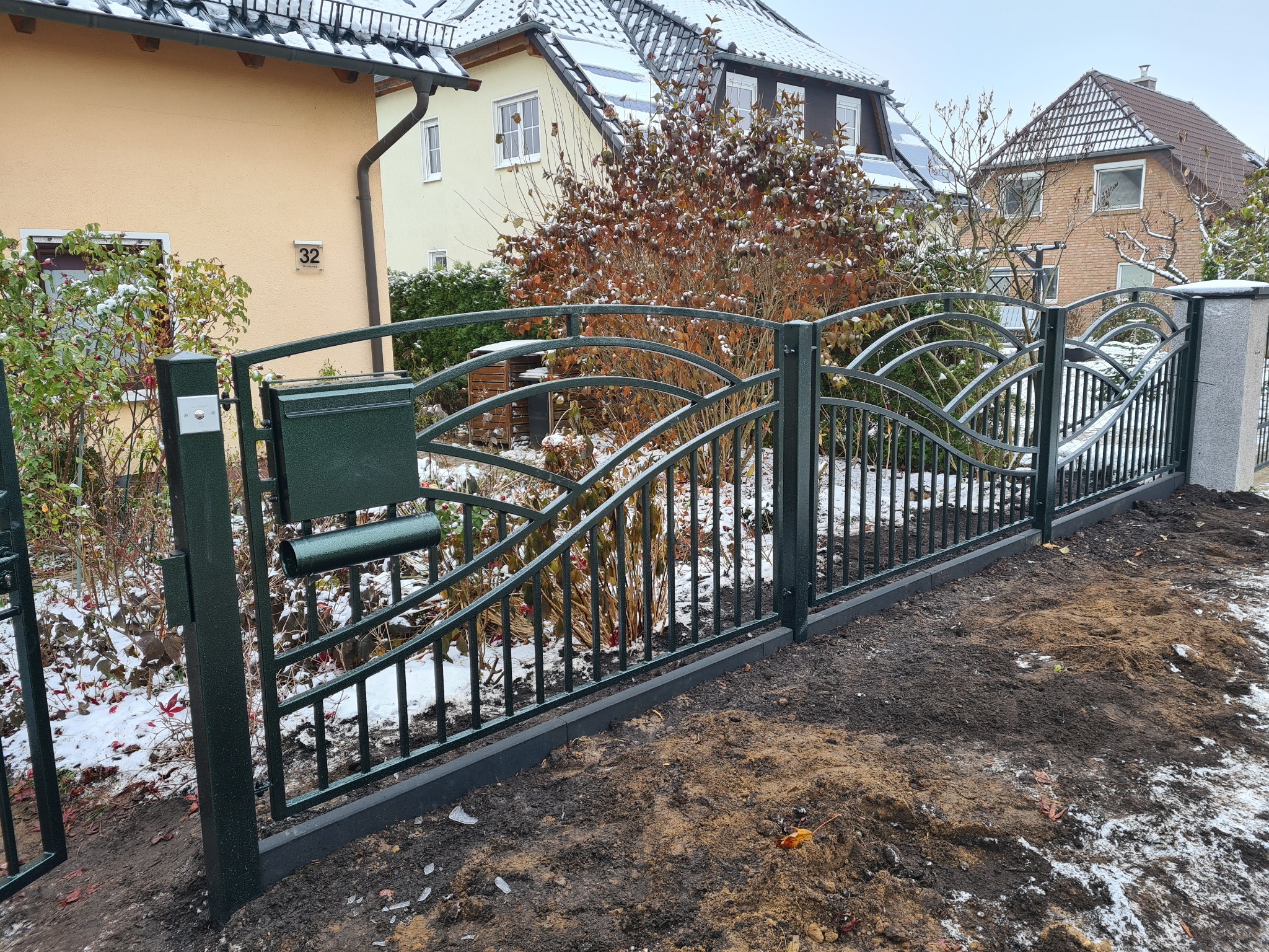 Nowoczesne, ciemnozielone ogrodzenie z metaloplastyki z przymocowaną skrzynką na listy, ustawione na świeżo przekopanym terenie z resztkami śniegu, na tle domów jednorodzinnych.