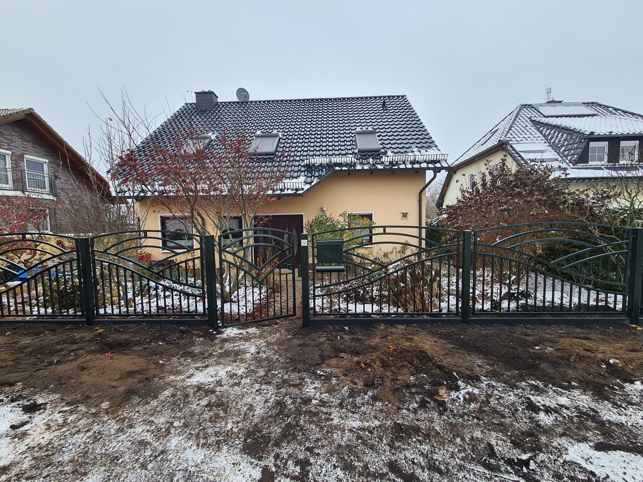 Kute ogrodzenie z otwartą bramą w kolorze ciemnej zieleni, z dekoracyjnymi łukami i pionowymi prętami, przed domem z dachówką pokrytą śniegiem w pochmurny dzień.