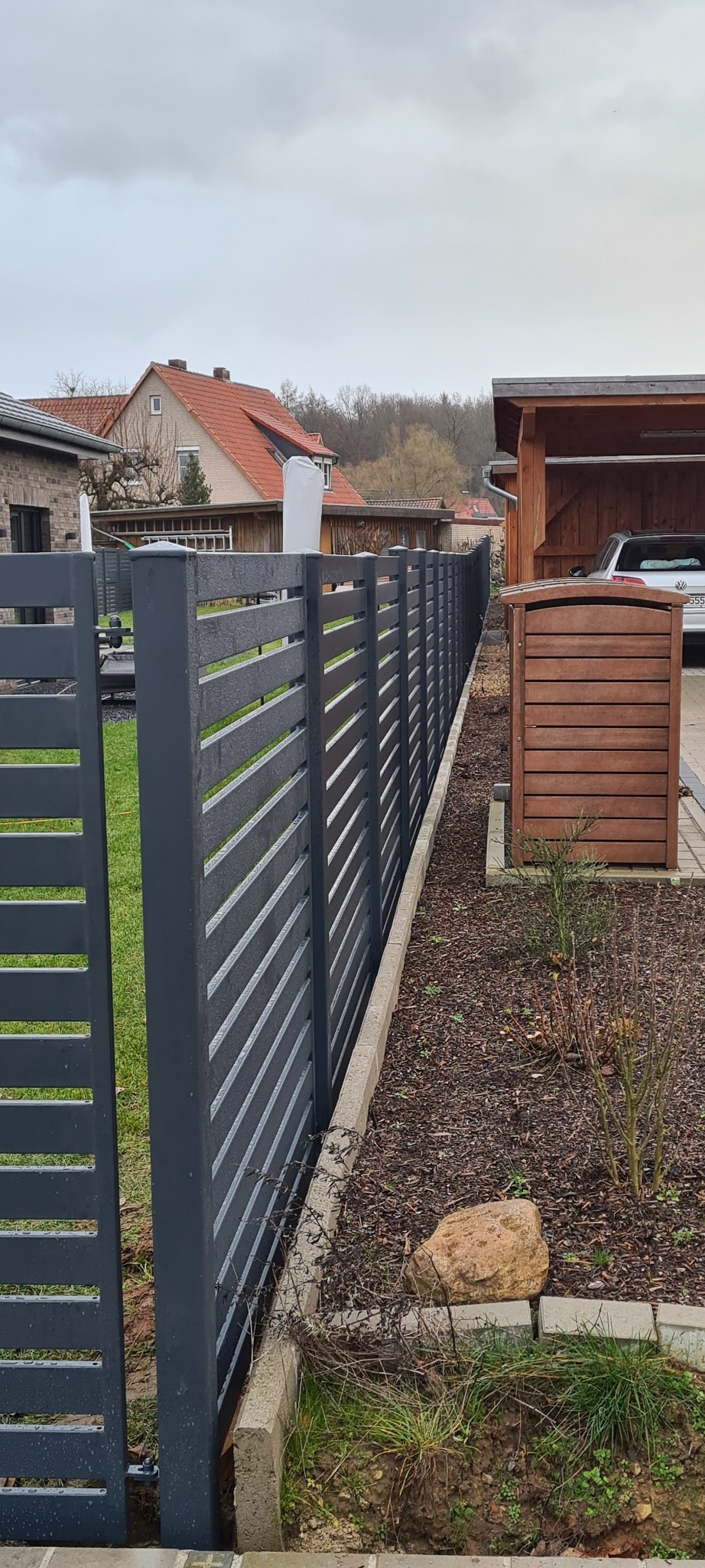 Nowoczesne, poziome, szare przęsła ogrodzeniowe z metalu, oddzielające posesję od sąsiada, widoczne z boku, z elementami dekoracyjnymi z kamienia wzdłuż podstawy i skrzynką na listy w tle.